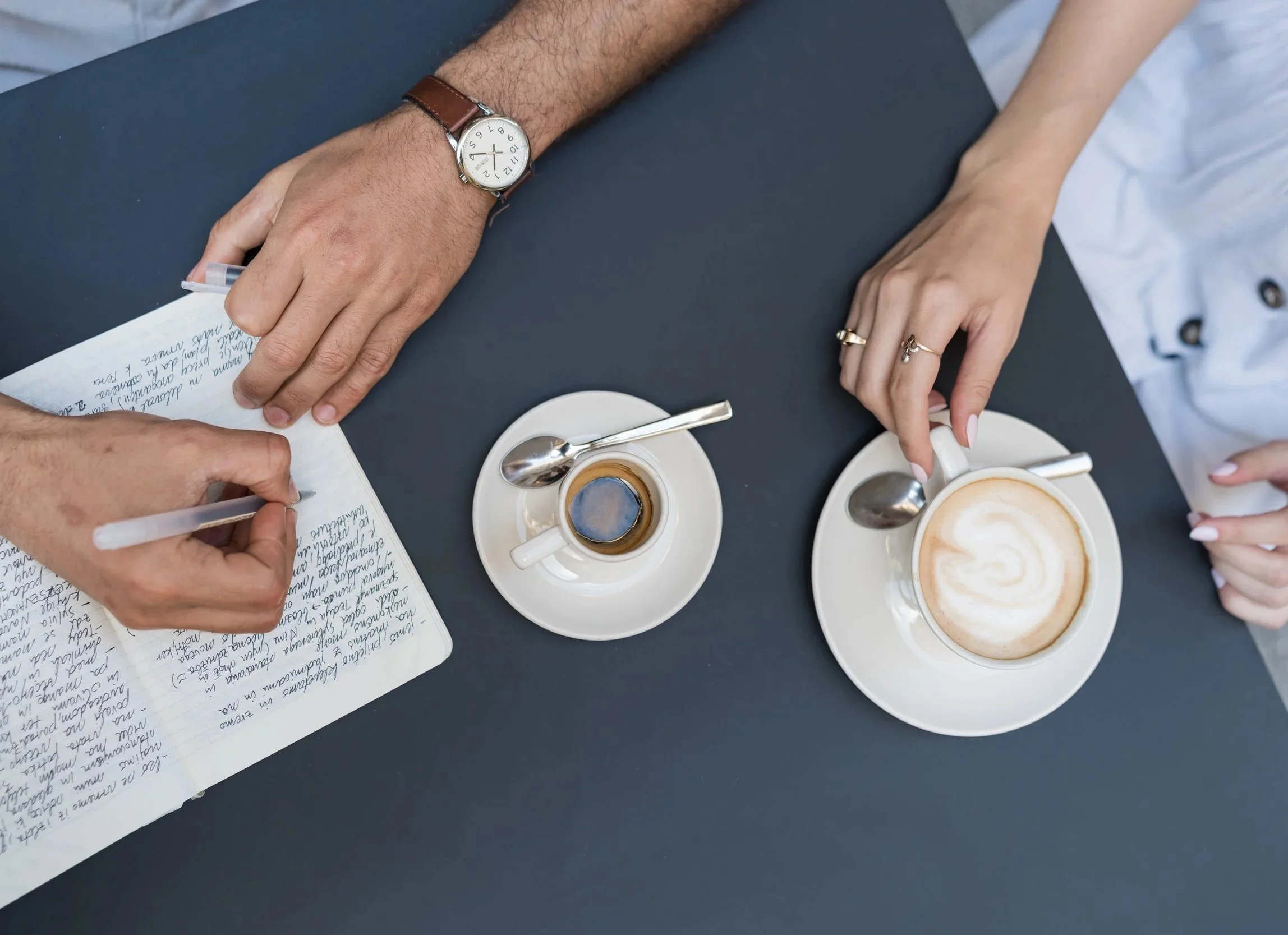 Image from above with hands on the table. One is taking notes in a journal. Two cups with saucers and spoons with an espresso and a latte