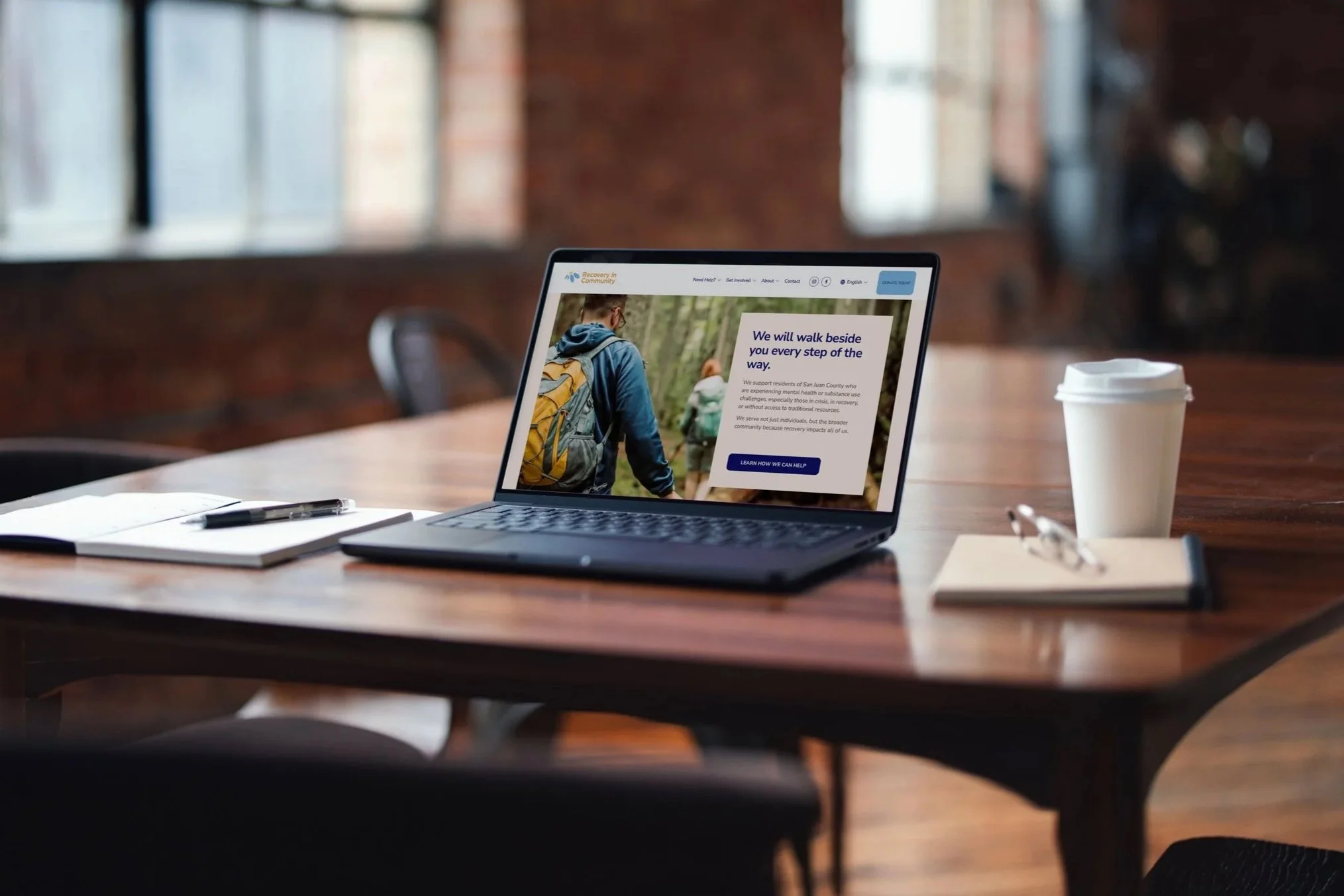 Laptop sitting on wooden deck with notepads, glasses, and coffee cup. Blurred windows in the background