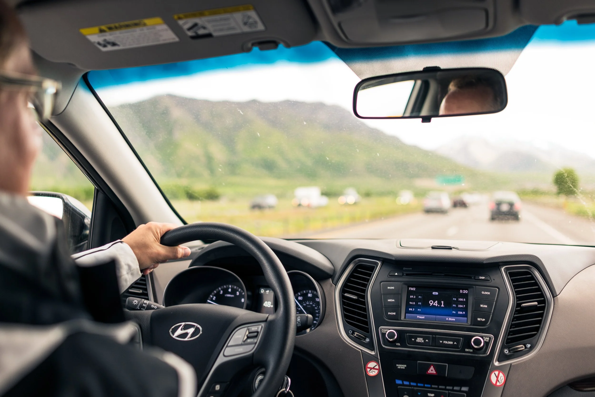 From back seat of car with partial view of driver wearing glasses on the left. Car is going down a two lane road in each direction with green hill in the background