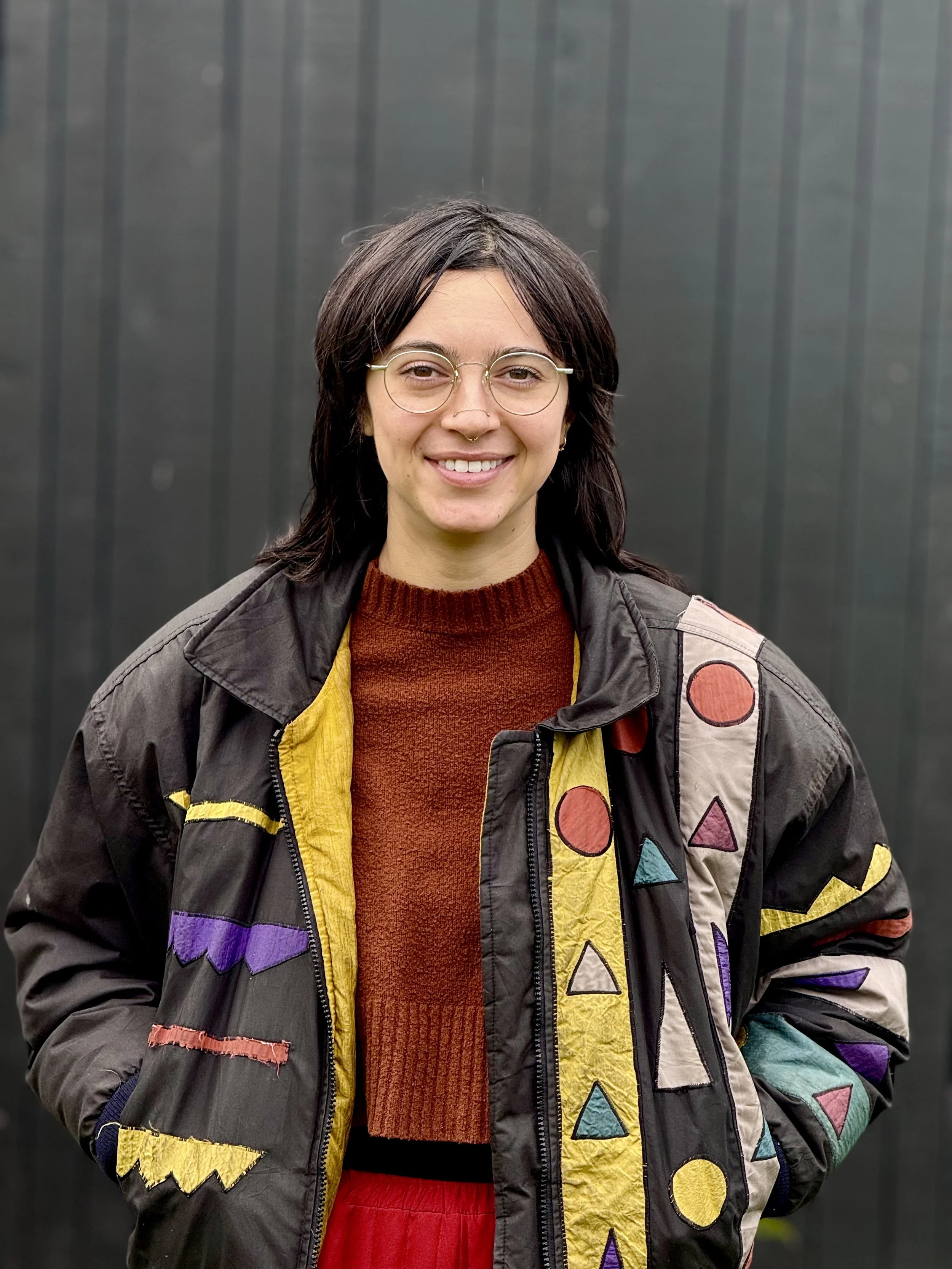 Portrait of a younger person with longish dark hair wearing round glasses, a black jacket with colorful geometric shapes and reddish-orange sweater in front of a dark gray background