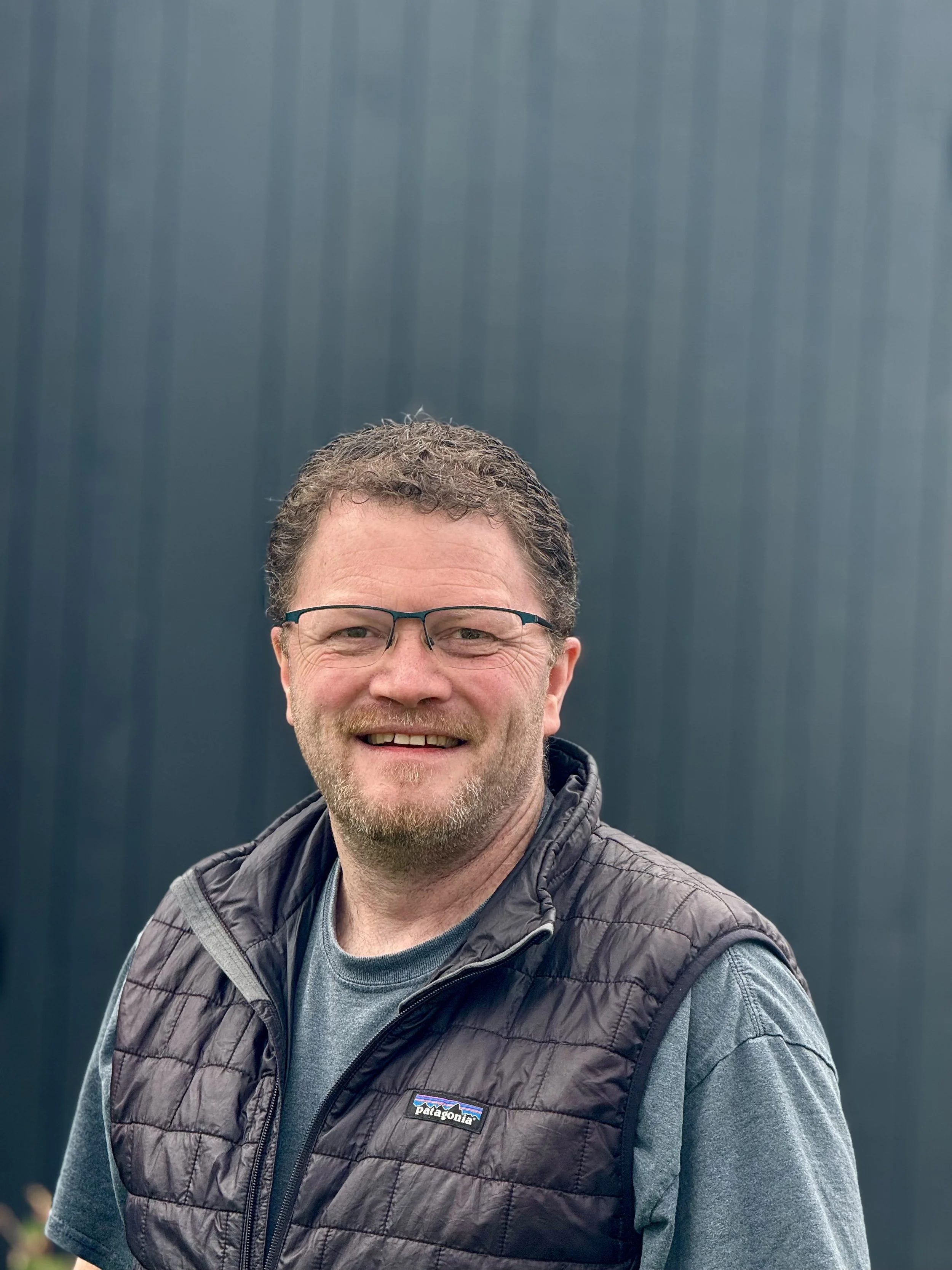 Portrait of a middle aged person with short reddish brown  hair wearing glasses with a black puffy vest and green t-shirt in front of a dark gray background