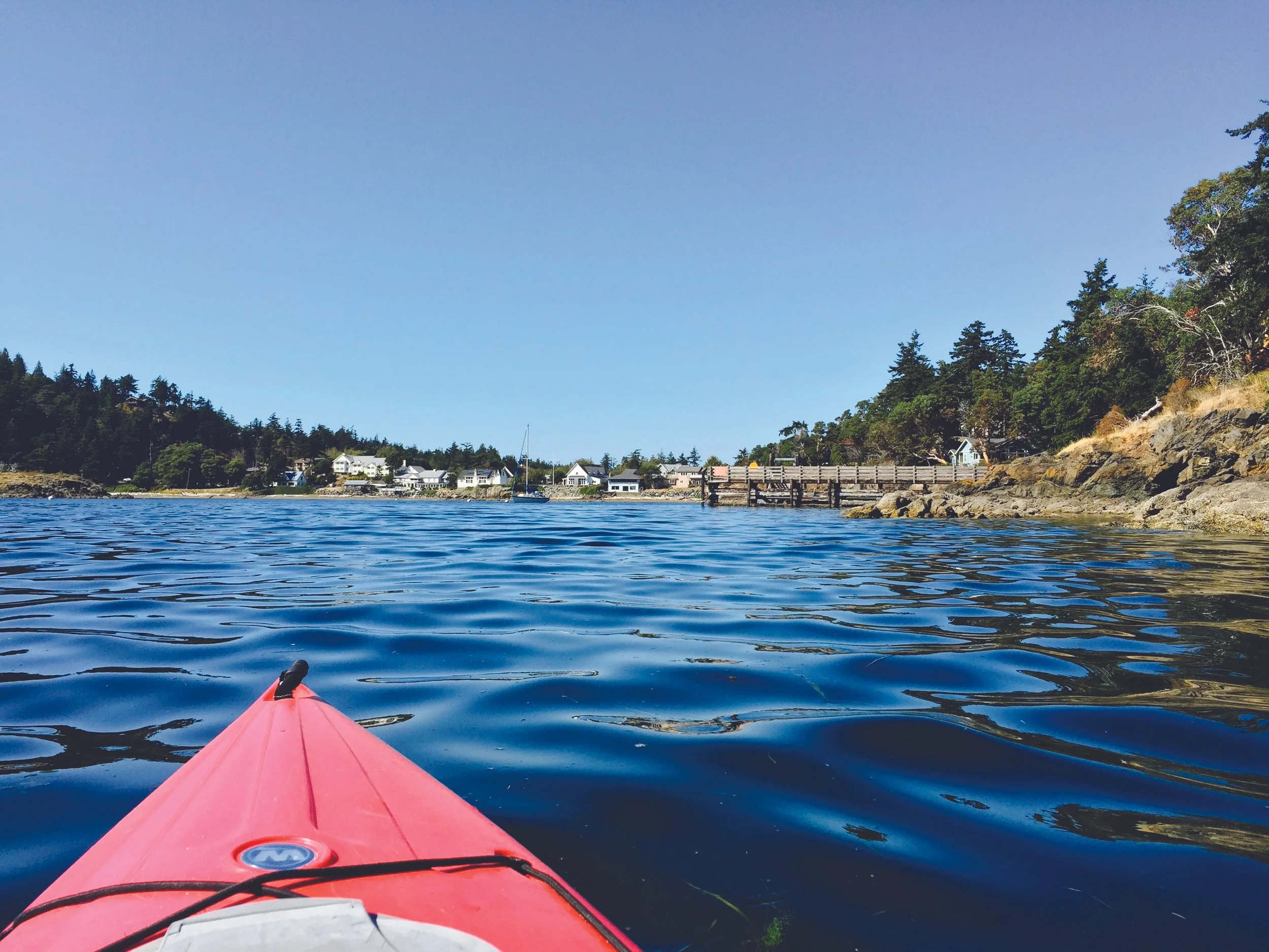 View point is from the front of a red kayak on the water. It looks towards a small town, Eastsound, with a pier in the foreground and conifer trees in beyond set against a blue sky