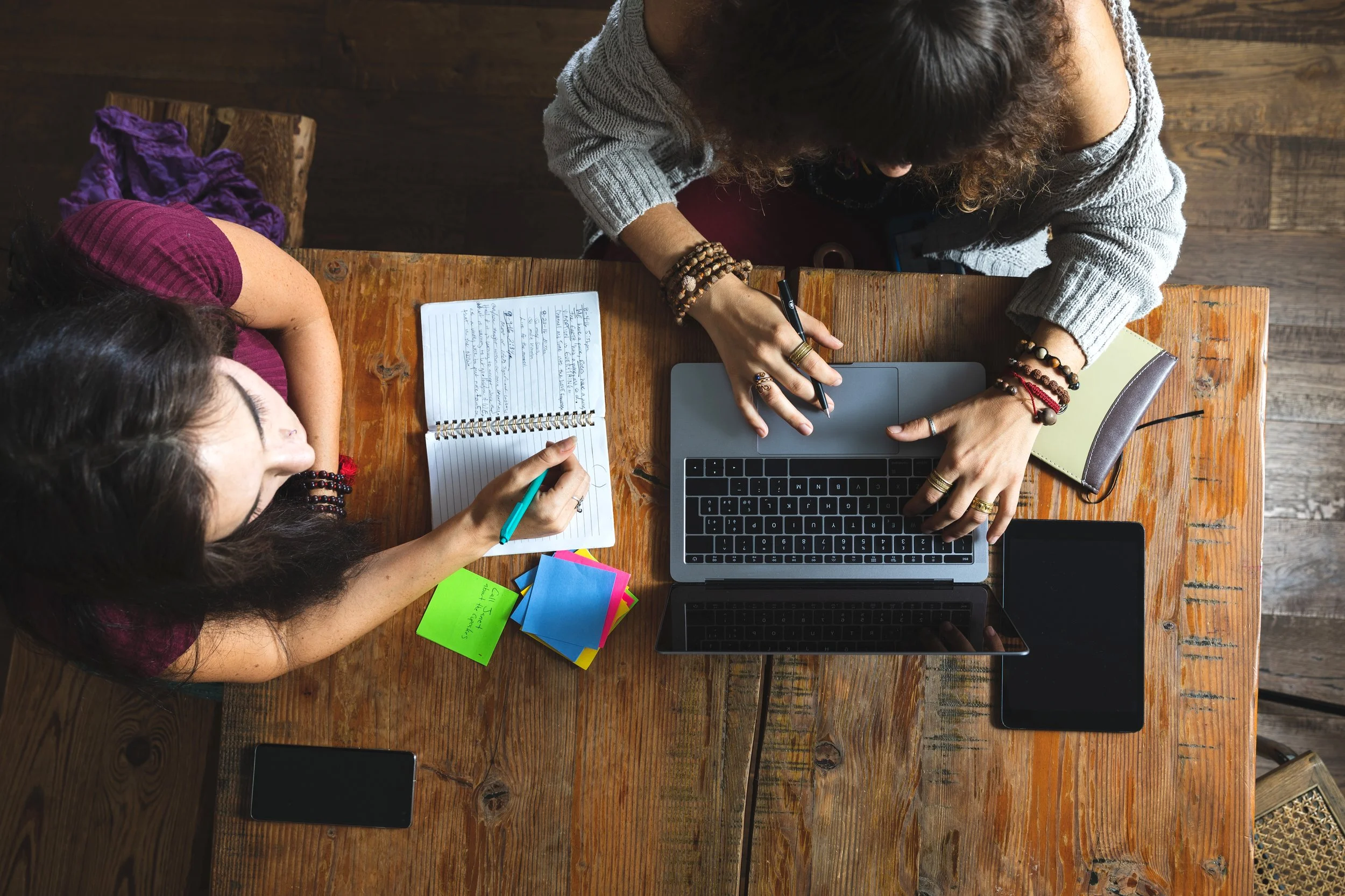 From above. Two people at wooden table. One person is taking notes in a journal with a phone and post its. The other is at a laptop wearing a grey sweater with a number of beaded bracelets on both wrists
