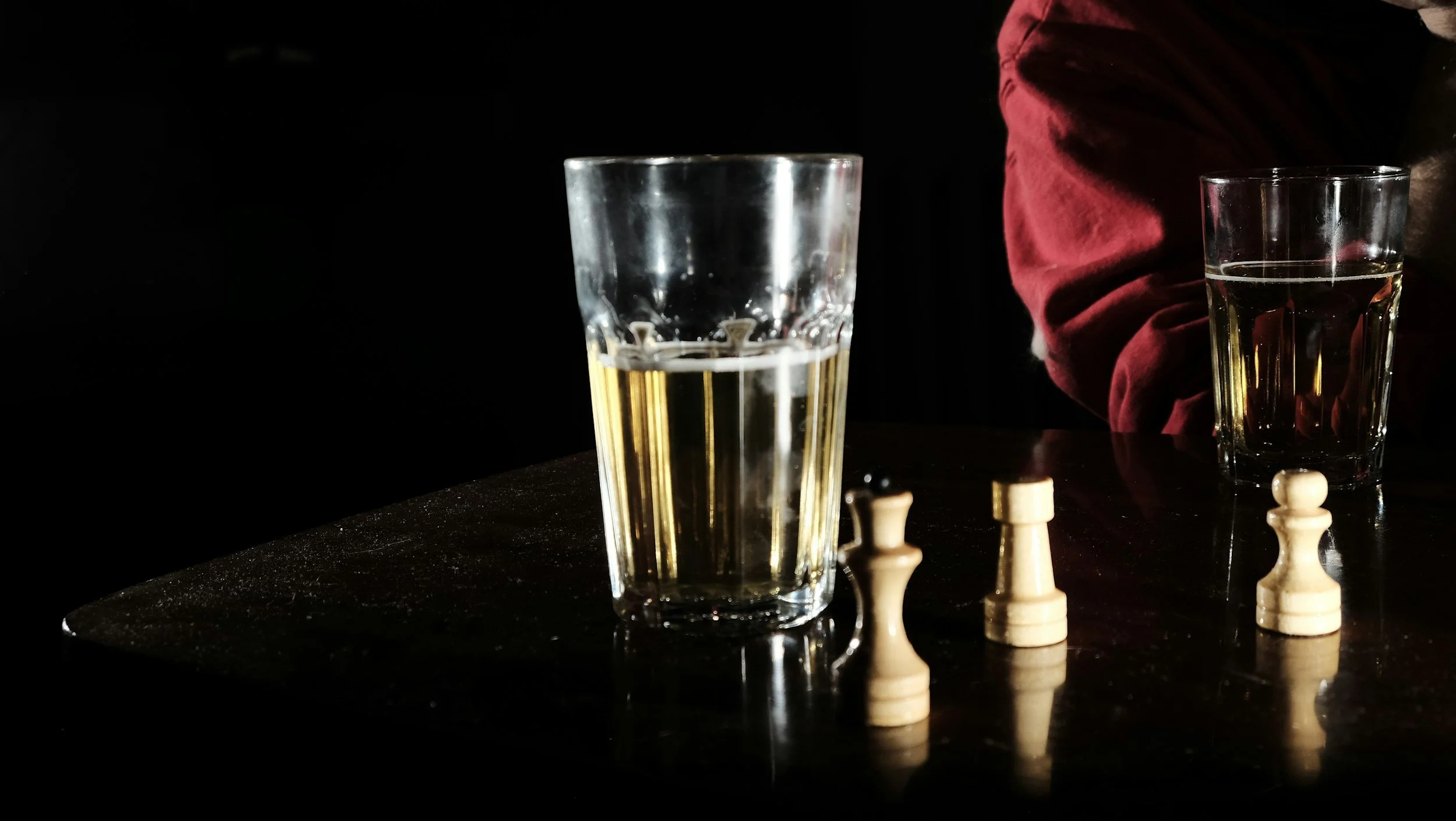 A glass of water, a glass of whiskey, and three chess pawns on a dark table, with a person wearing a red shirt partially visible in the background.