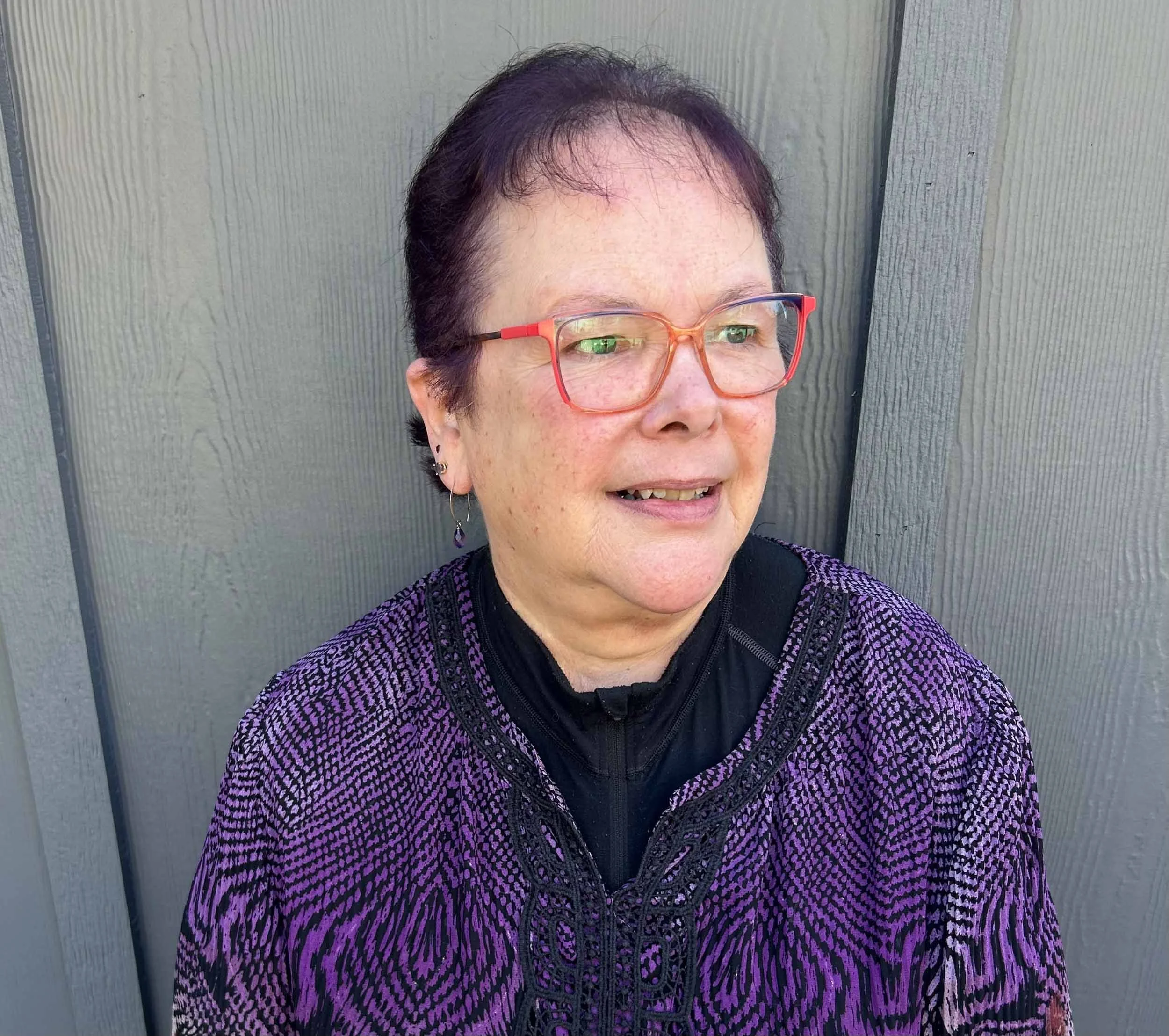 A woman with short dark hair, wearing red-framed glasses, earrings, and a purple patterned jacket, standing outdoors against a gray wooden fence, looking to the right.