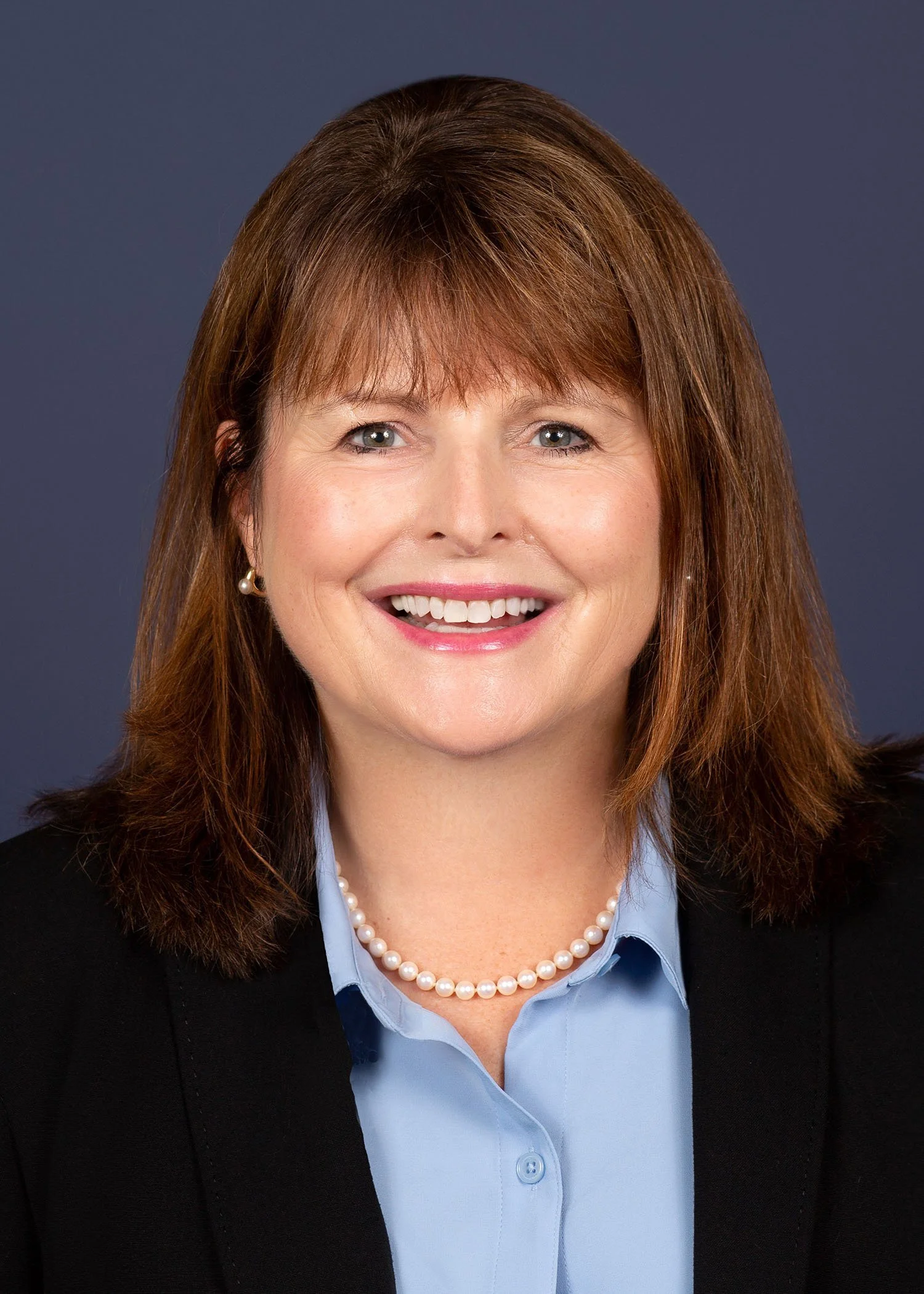 A professional woman with shoulder-length brown hair, wearing a black blazer, light blue shirt, pearl necklace, and pearl earrings, smiling against a dark background.