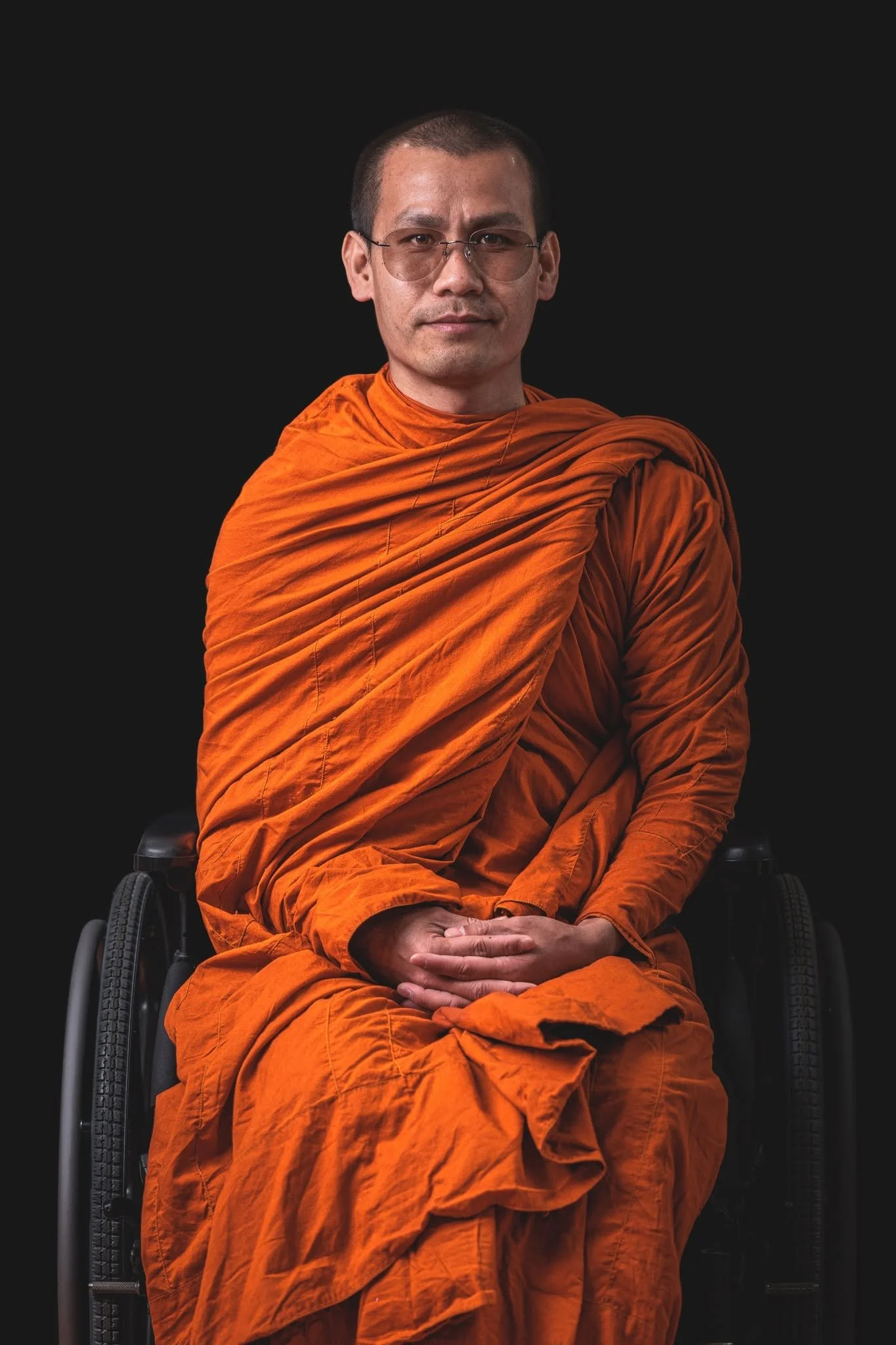 A man in orange robes sitting in a wheelchair against a black background.