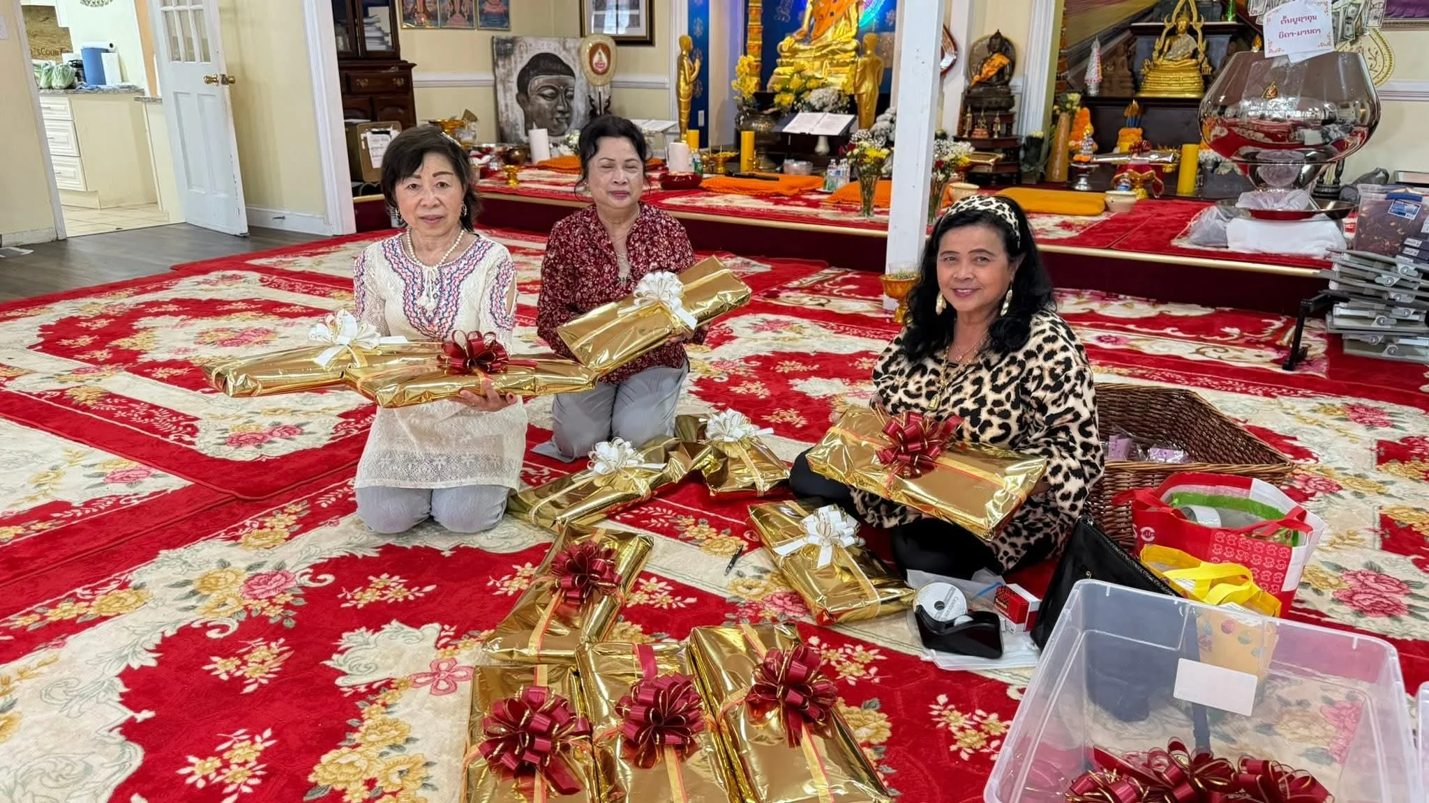 Three women sitting on a red and cream floral patterned rug, holding wrapped gifts with gold paper and decorative bows, in a room with religious and decorative items, and a table with more presents and supplies.