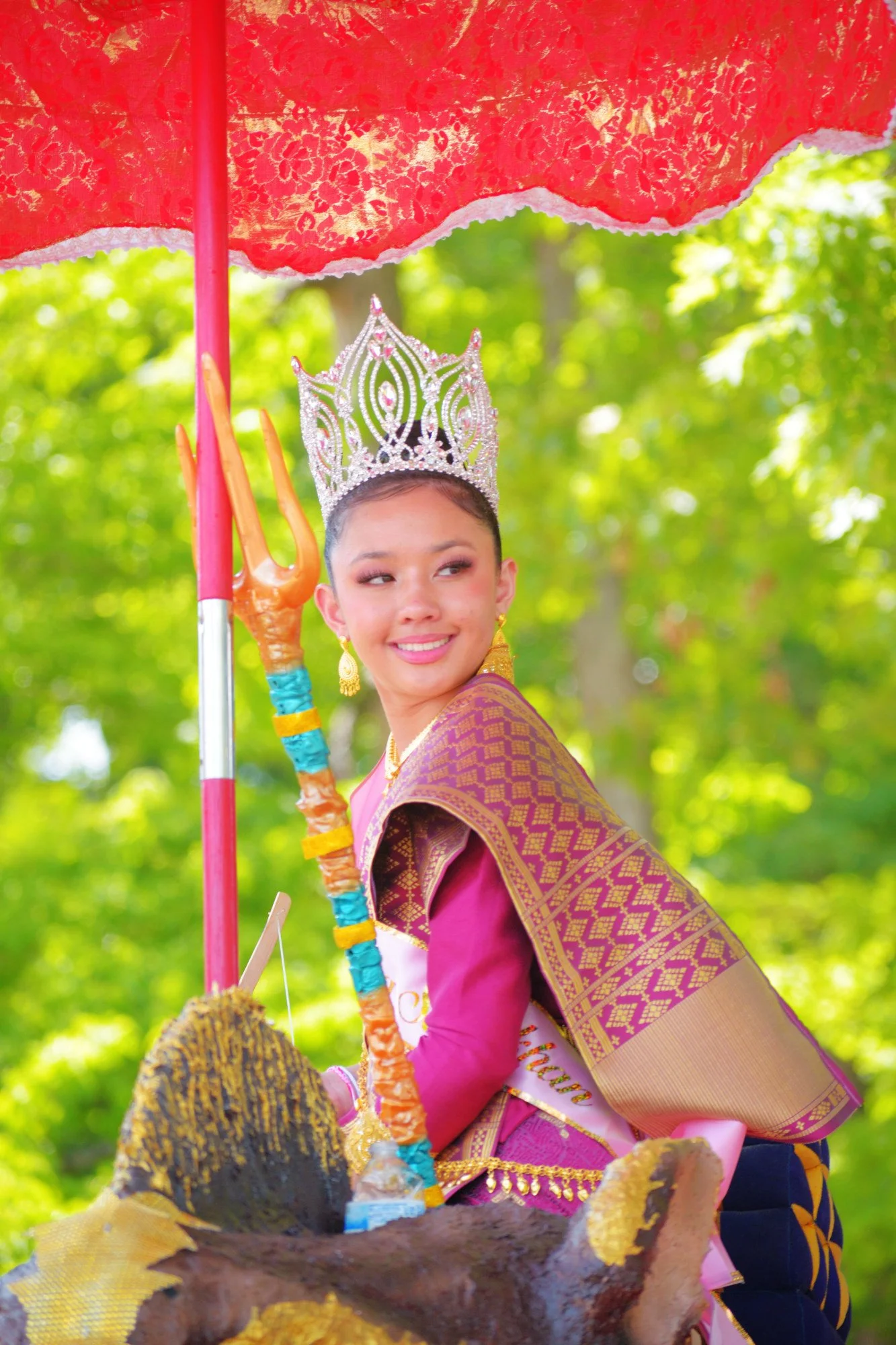 A woman dressed in colorful traditional attire with a crown on her head, holding a trident, standing outdoors under a red umbrella.