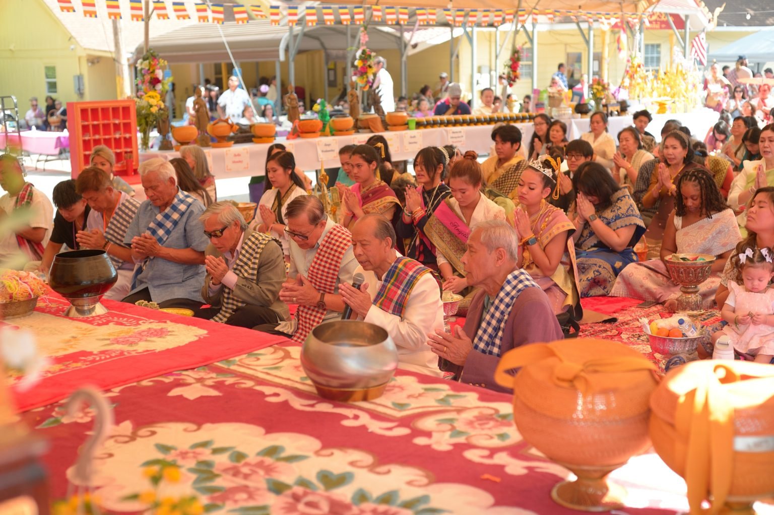 Group of people participating in a religious or cultural ceremony outdoors under a large tent, with some people wearing traditional clothing and sitting with hands in prayer.
