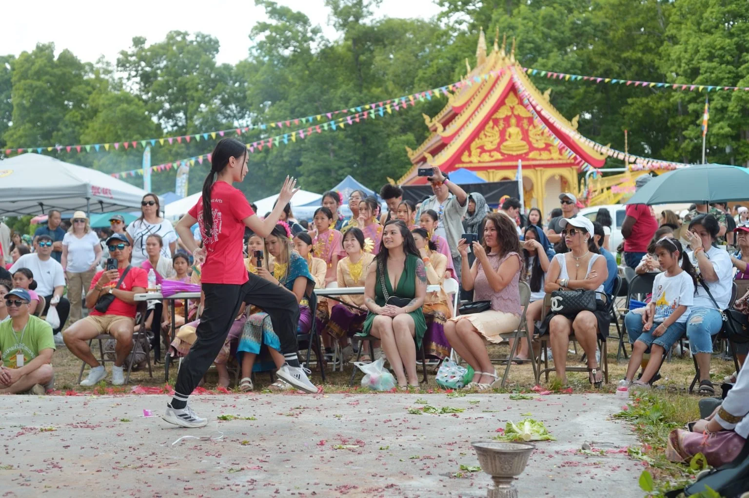 A person in a red shirt appears to be performing or demonstrating in front of an outdoor audience at a cultural festival. The audience is seated and taking photos or watching. There are colorful tents, flags, and a decorated temple structure in the background, suggesting a festive, cultural event in a park.