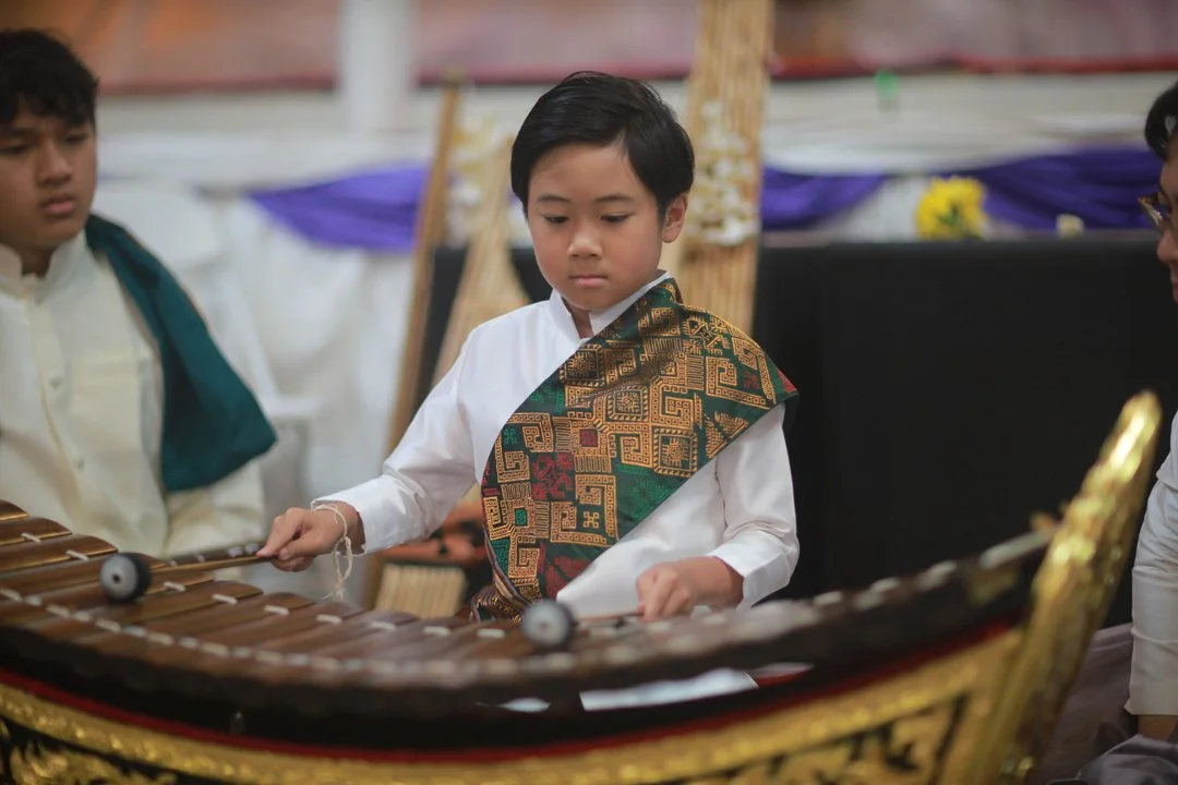 Young boy playing a traditional wooden xylophone during a cultural event, wearing a white shirt and a colorful patterned sash.