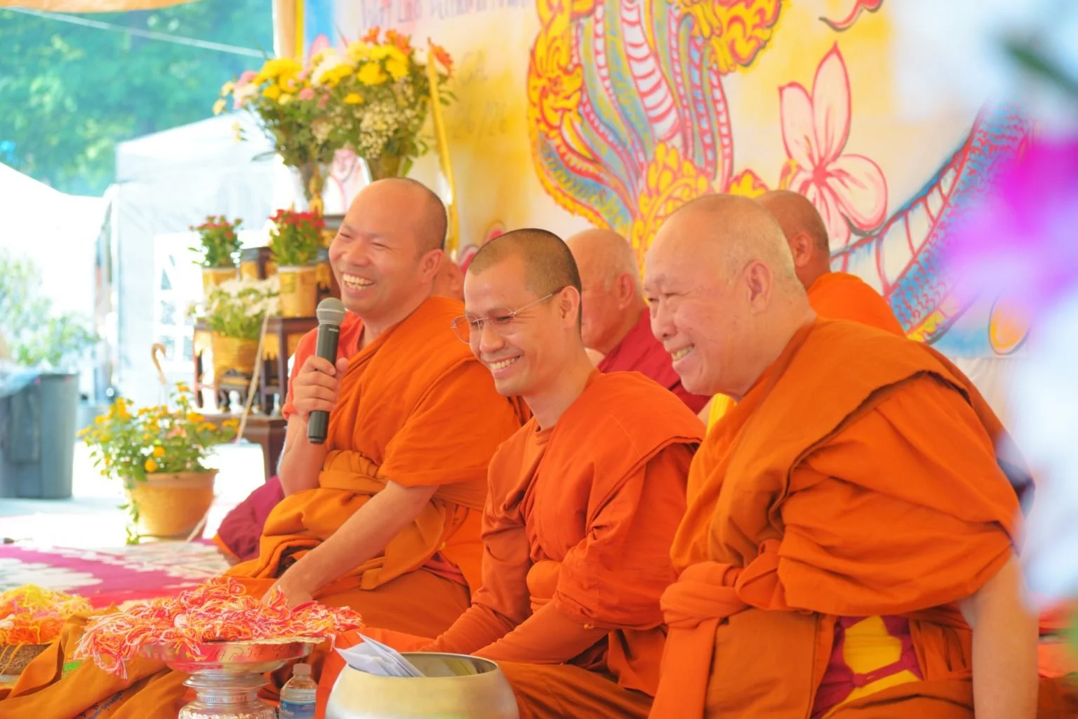 Group of Buddhist monks in orange robes sitting together at a celebration, with one holding a microphone and everyone smiling, decorated with colorful banners and flowers.