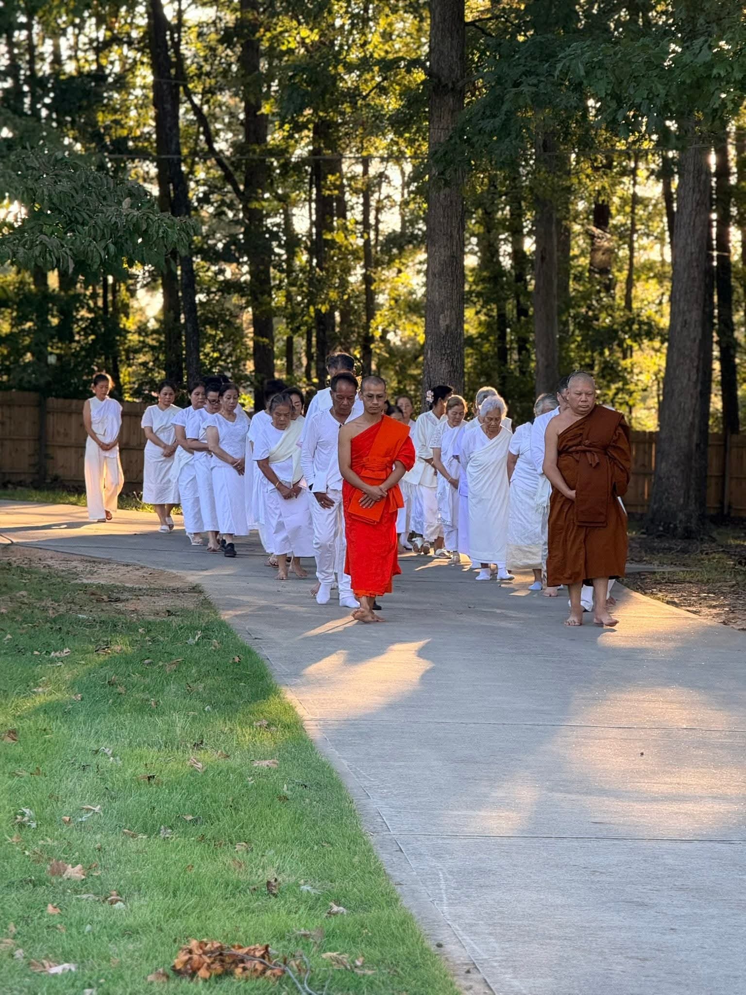 A group of Buddhist monks and nuns, dressed in white and orange robes, walking in a line on a paved path in a wooded area.
