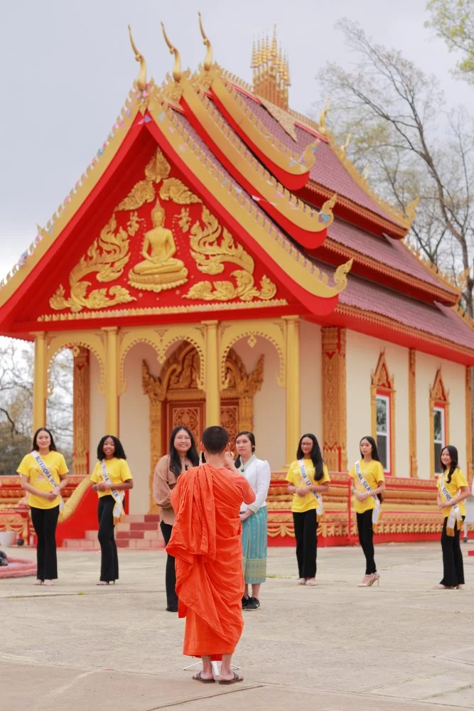 A Buddhist monk in orange robes standing in front of a group of women, some wearing yellow shirts with sashes, outside a traditional Thai temple with ornate red and gold decorations and a golden Buddha statue on the gable.