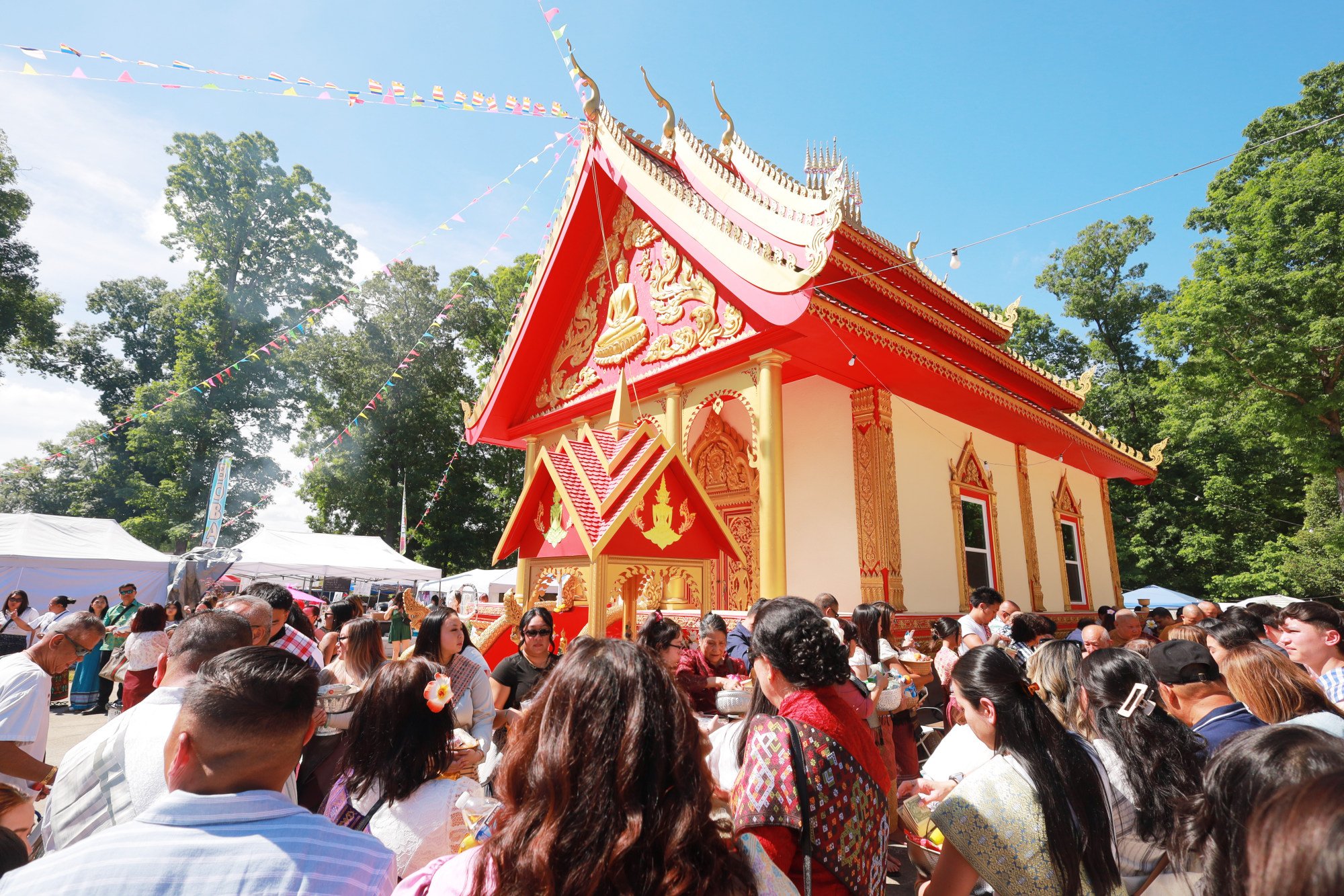 A crowded outdoor scene at a temple festival with people gathered around a decorated temple building under a blue sky, surrounded by green trees and colorful bunting.