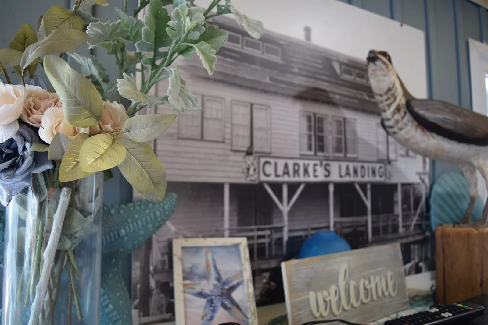 Decorative scene with a glass vase of artificial flowers in the foreground, a picture of a sailboat, a starfish, a wooden "welcome" sign, a painted sign that reads "Clarke's Landing," a mounted decorative fish, a carved wooden whale figure, and surfboards in the background, suggesting a coastal or beach theme.