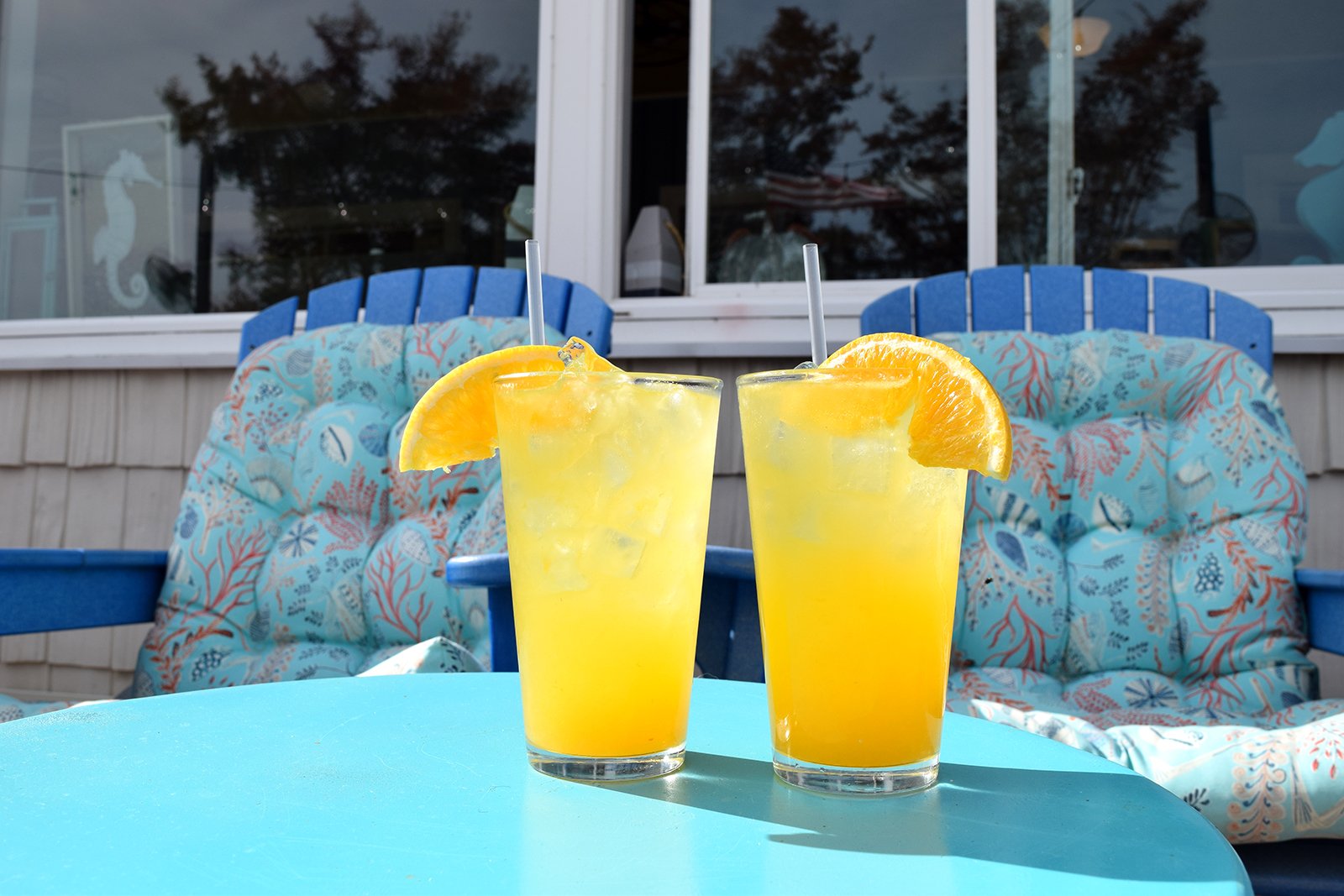Two glasses of yellow-orange cocktails with orange wedge garnishes on a blue table, outdoor patio with blue chairs and floral cushions in the background.