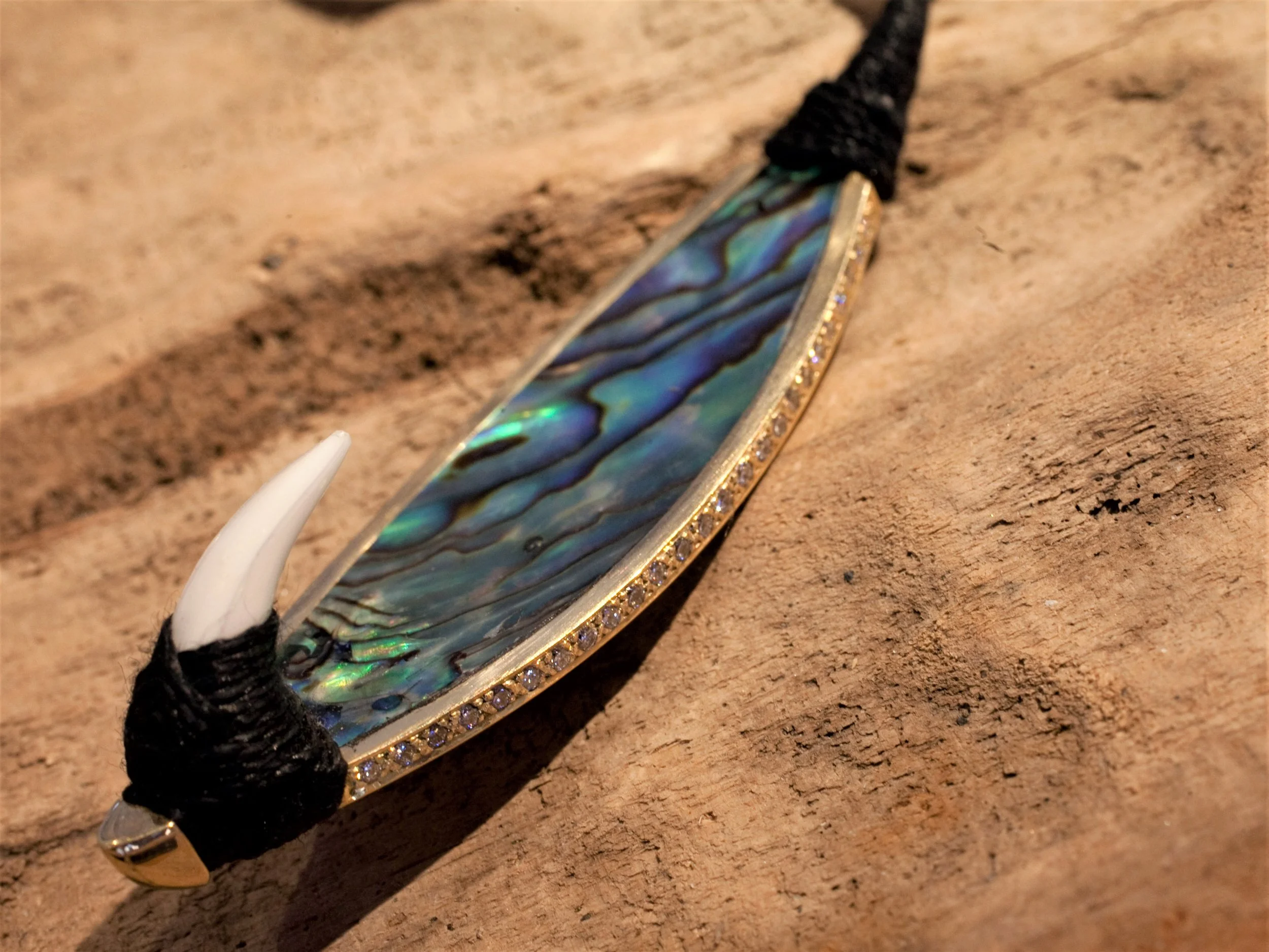 A necklace with an iridescent, shell-like pendant adorned with small gemstones along its border, attached to a black cord with white claw-like end caps, lying on a wooden surface.