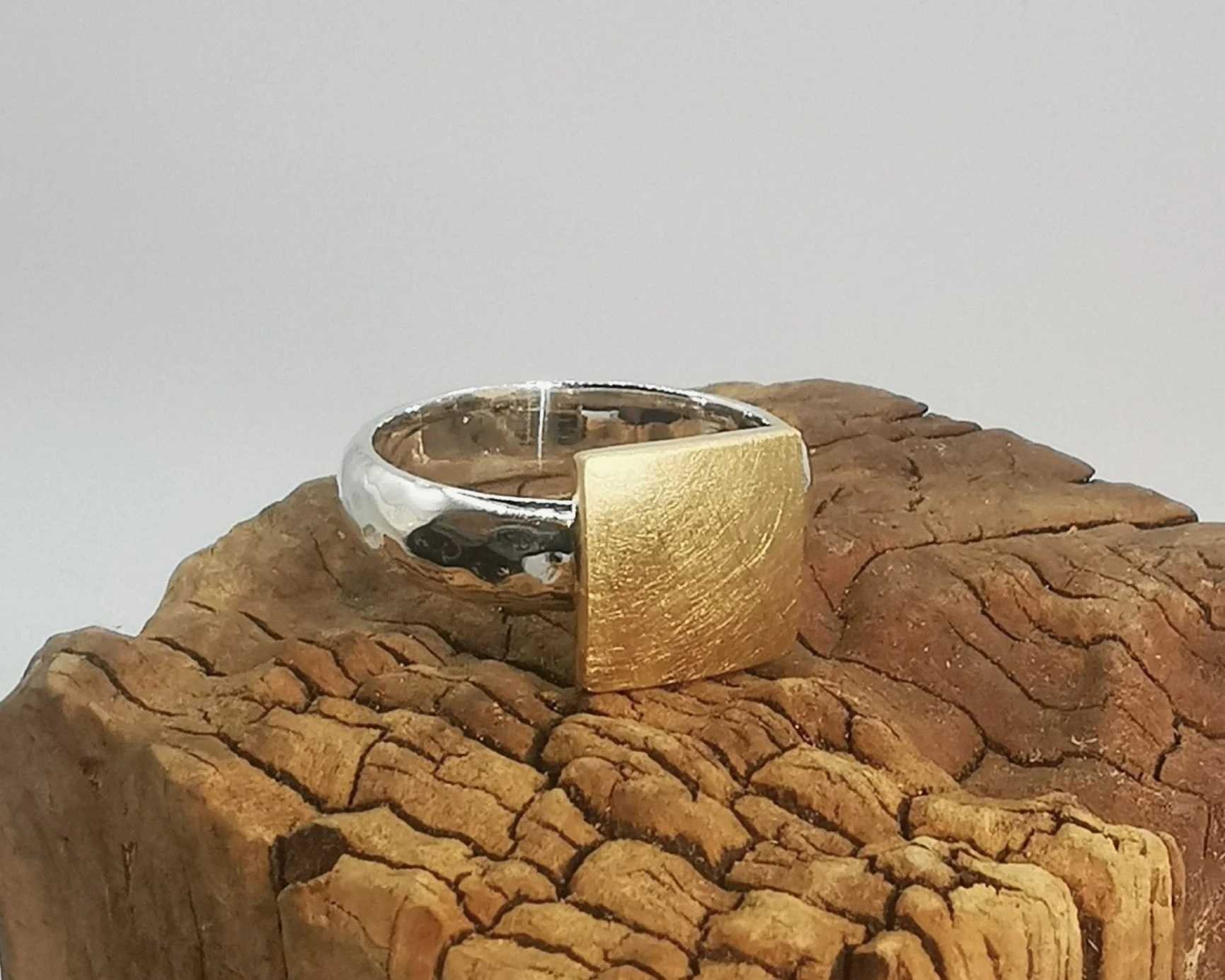 A silver ring with a wide, flat gold-colored rectangular face, resting on a textured piece of wood.
