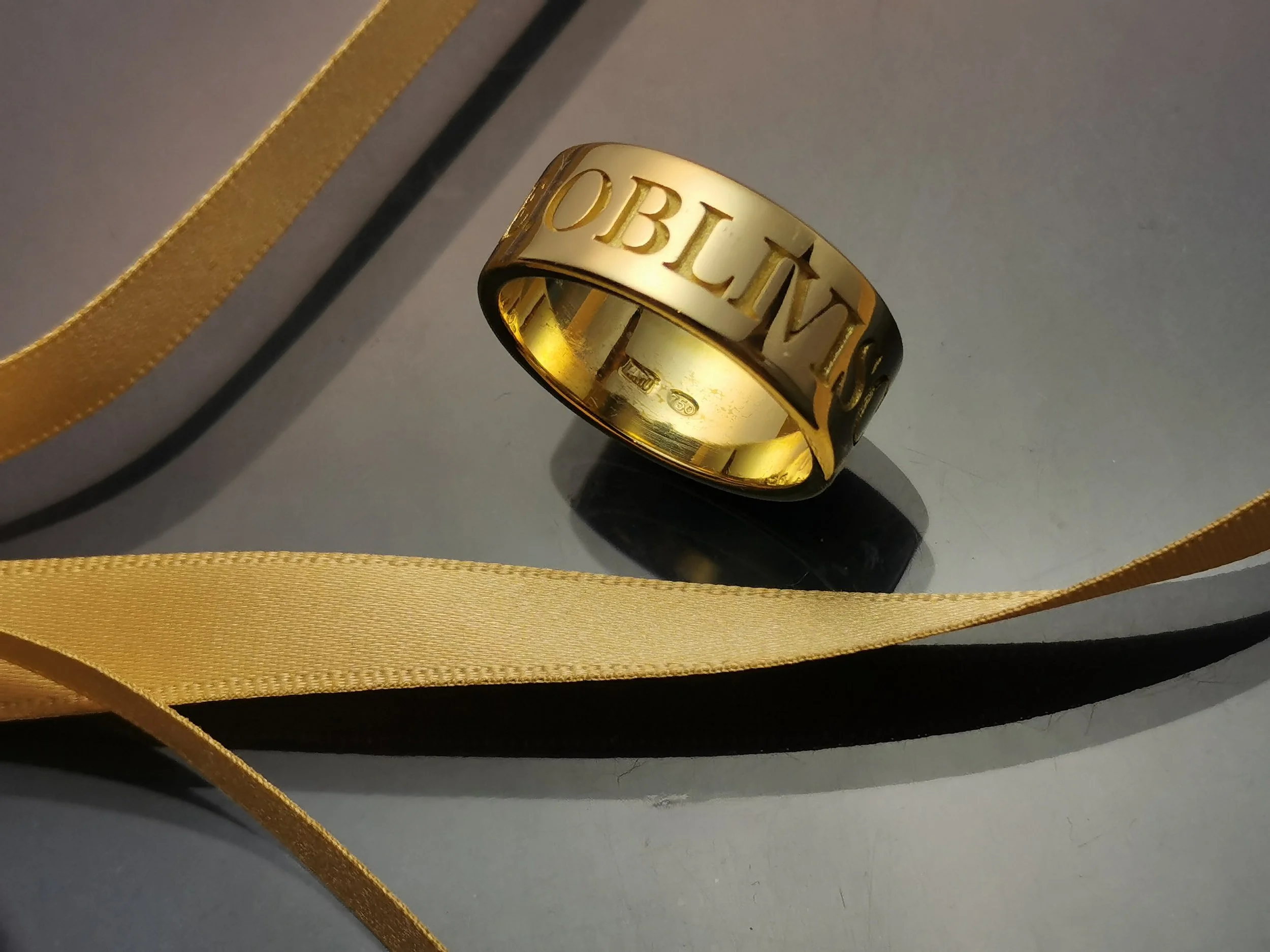 A gold ring with the word 'OBLIVION' engraved on it, placed on a reflective surface, with gold and gold-silver ribbons surrounding it.