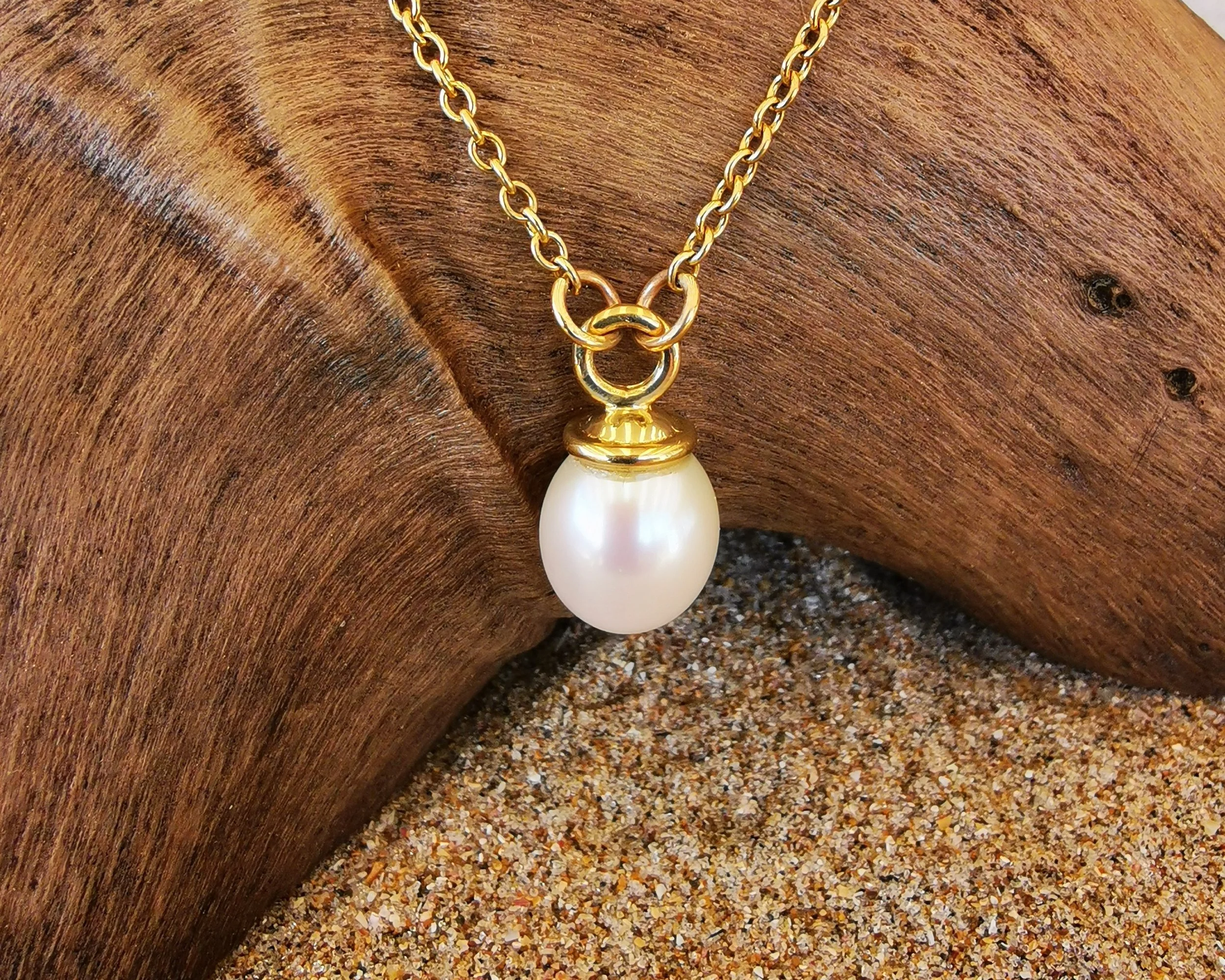 A pearl pendant necklace with a gold chain and clasp, hanging from a curved piece of wood on sandy ground.