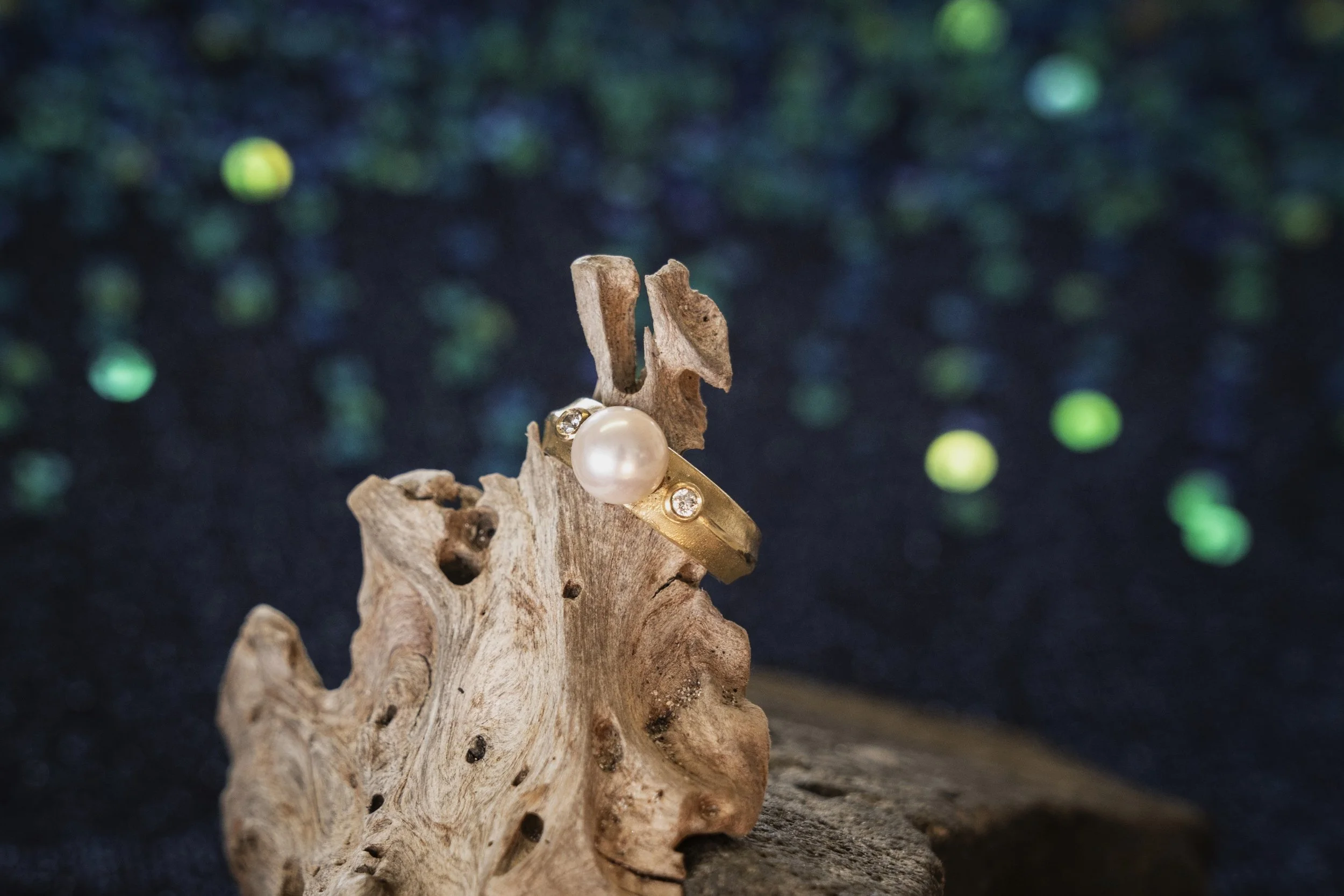 A piece of driftwood displays a pearl ring and diamond earrings against a dark, bokeh background with green dots.