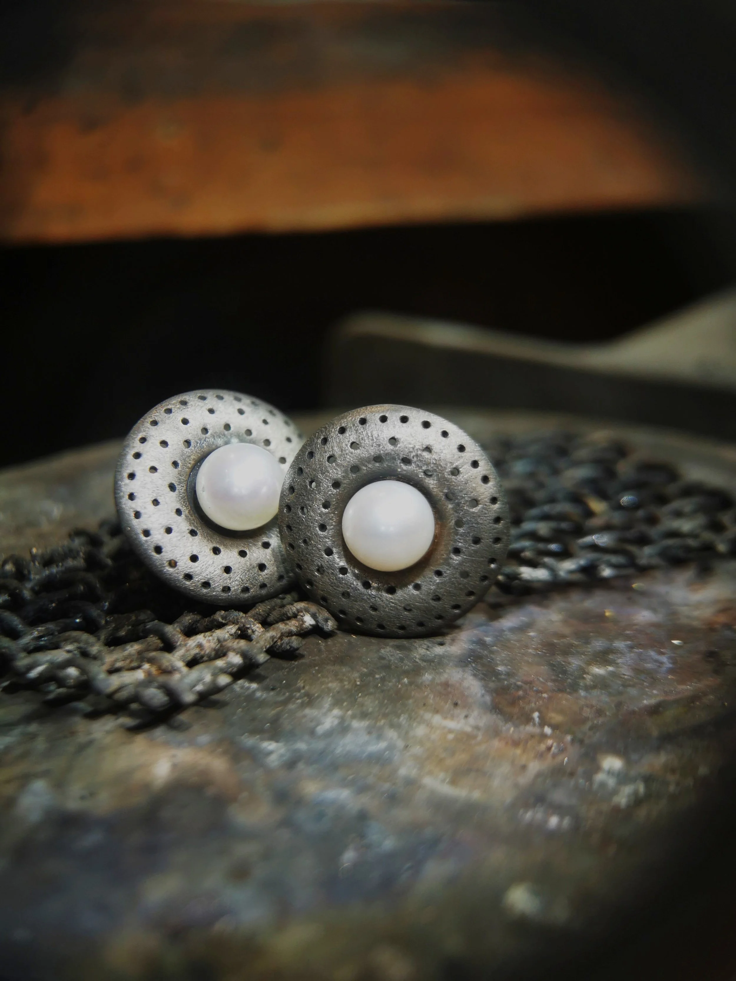 Pair of gray circular earrings with white pearl centers and small perforations, placed on a textured surface.