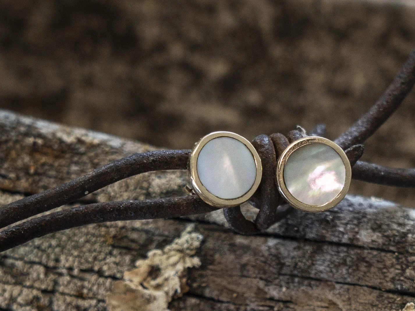 Close-up of earrings with round, iridescent stones in metal settings, resting on rusty barbed wire on a weathered wooden surface.