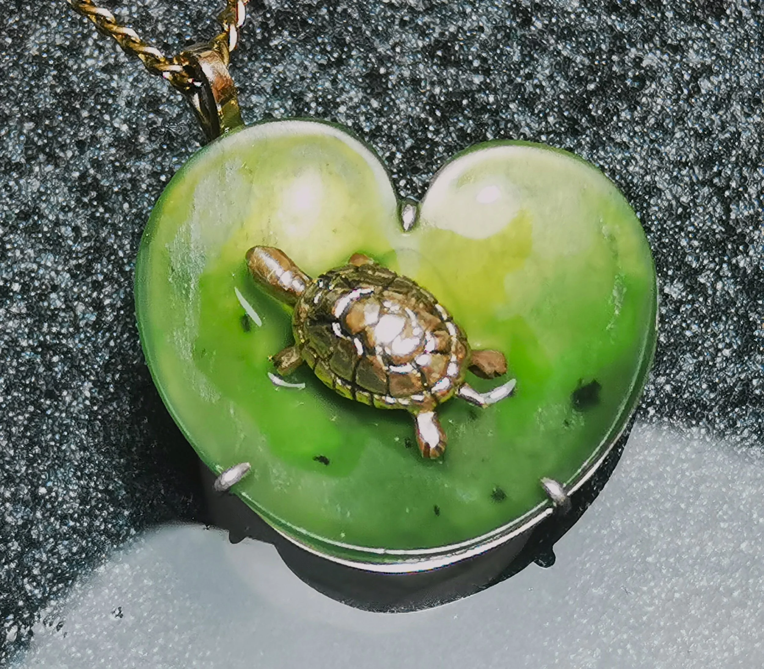 A small turtle on a green, heart-shaped, leaf-shaped object with a chain around it, on a textured black surface.