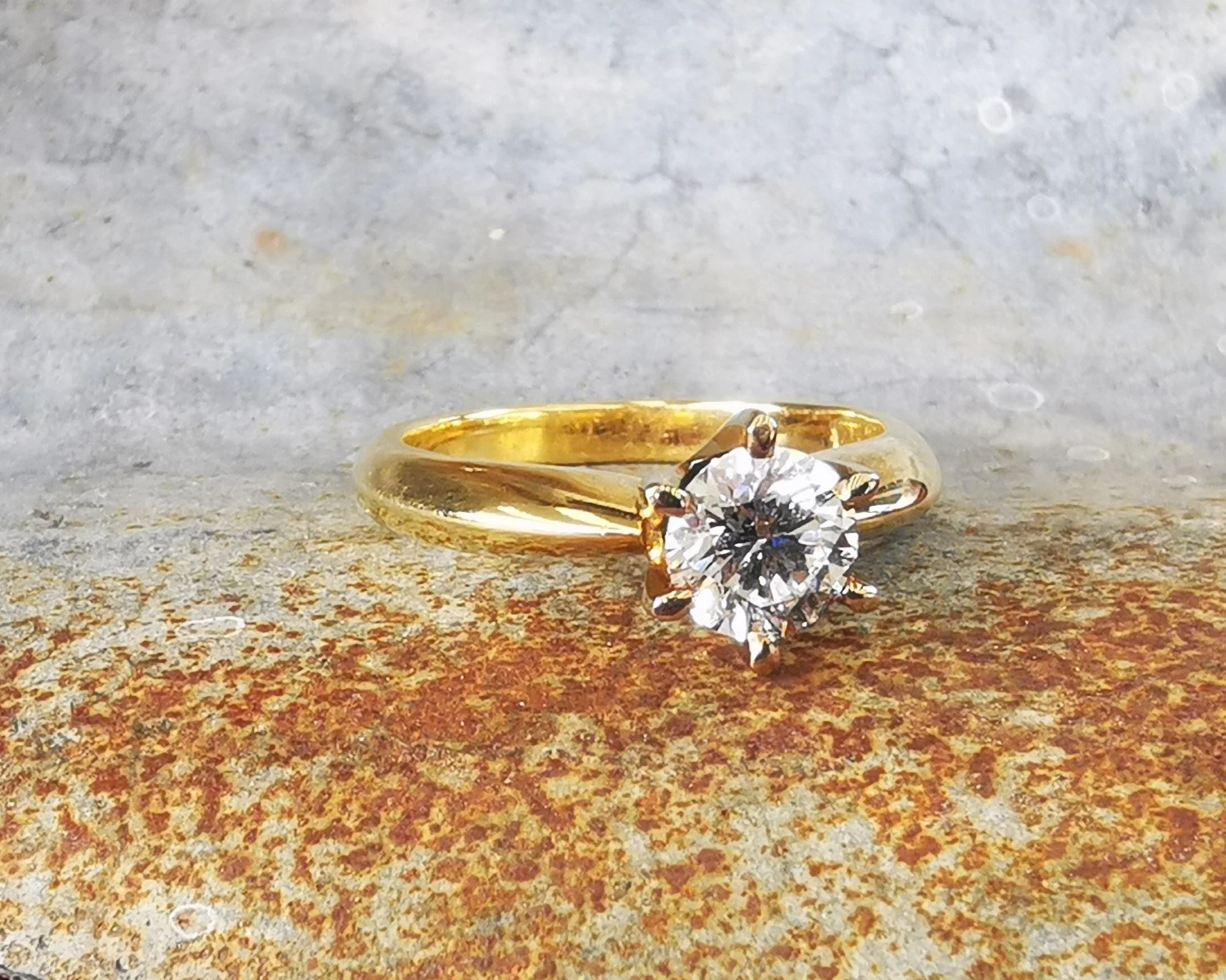 A gold engagement ring with a large round diamond solitaire set in a prong setting, resting on a rusted metal surface.
