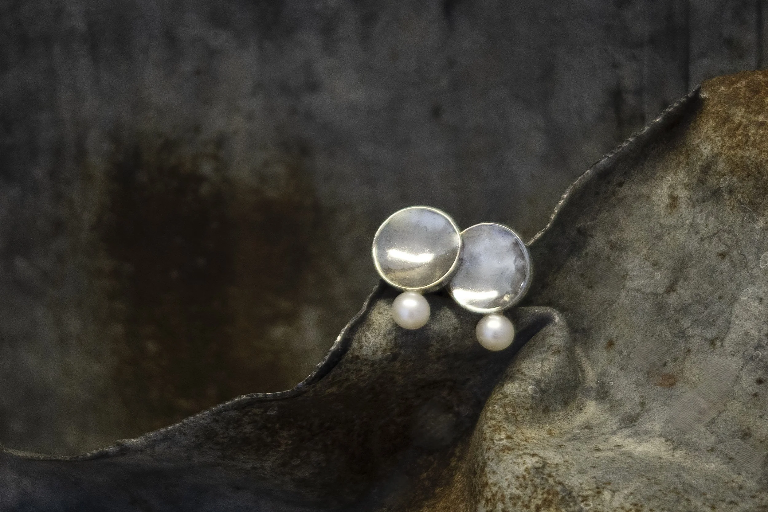 A pair of pearl earrings with transparent round stones set in silver, resting on a textured, rust-colored metal surface.