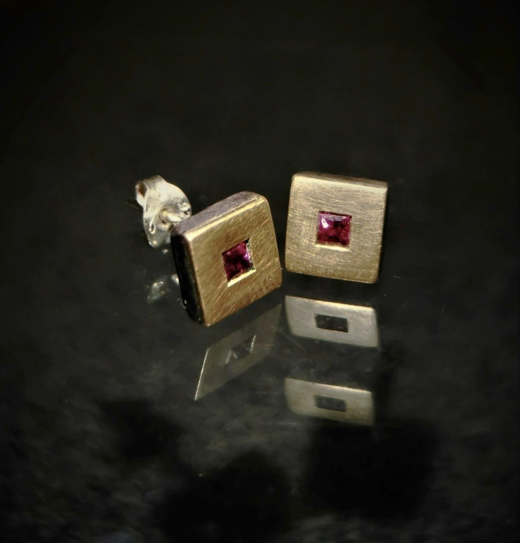 Gold-colored square earrings with purple gemstones in the center, placed on a reflective black surface.