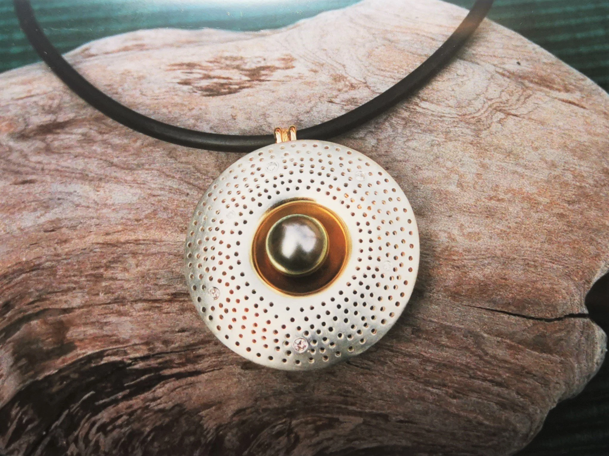 A circular pendant with a central black pearl surrounded by gold, dotted with small holes, hanging on a black cord, placed on a piece of textured wood.