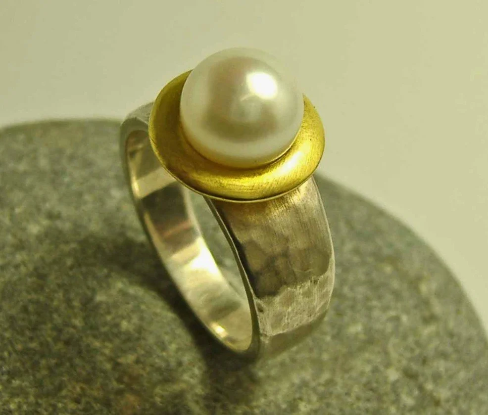 Silver ring with a gold bezel setting, featuring a large white pearl on top, placed on a gray stone surface.