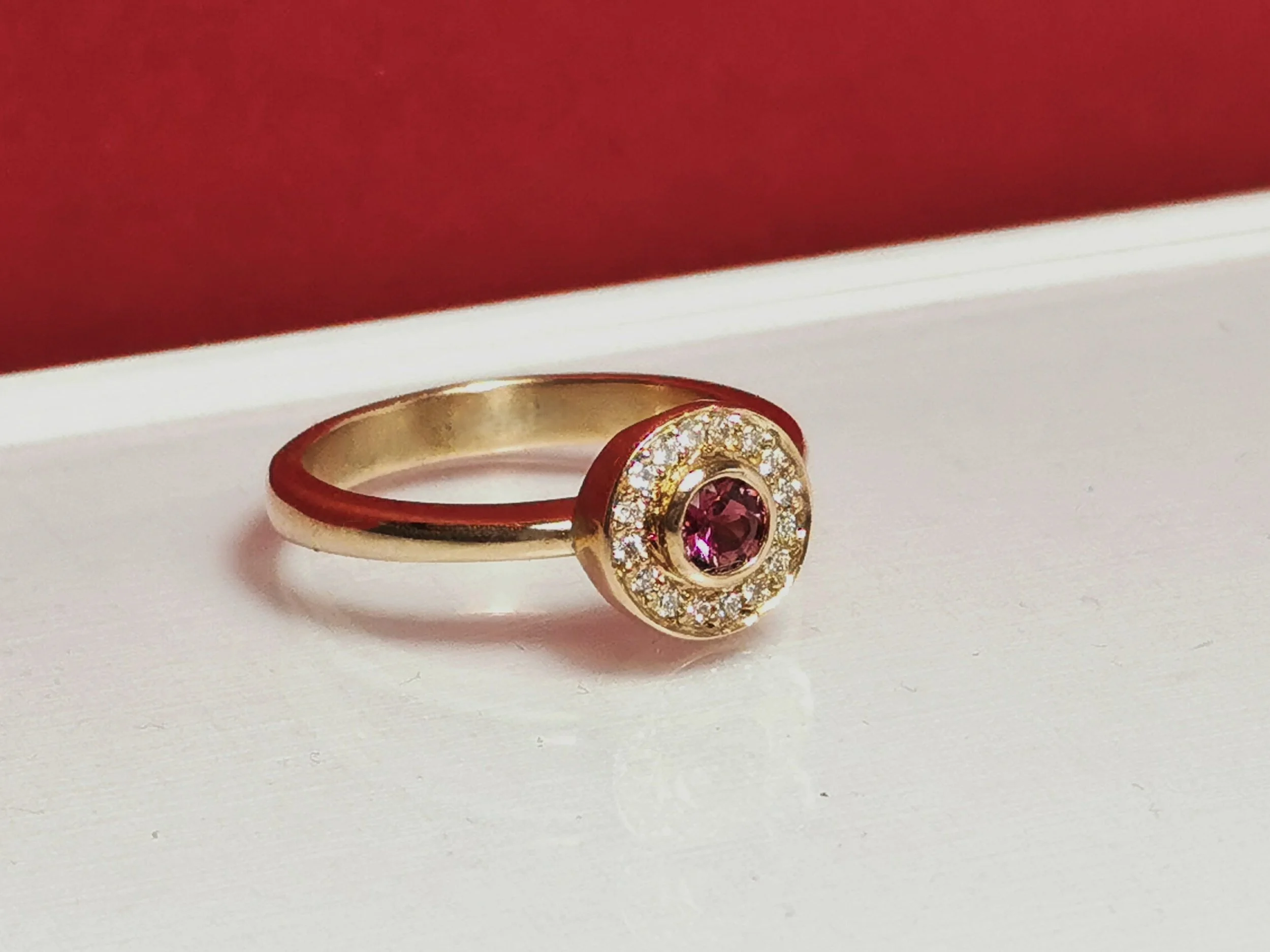 Gold ring with a pink gemstone center surrounded by small diamonds, placed on a white surface with a red background.