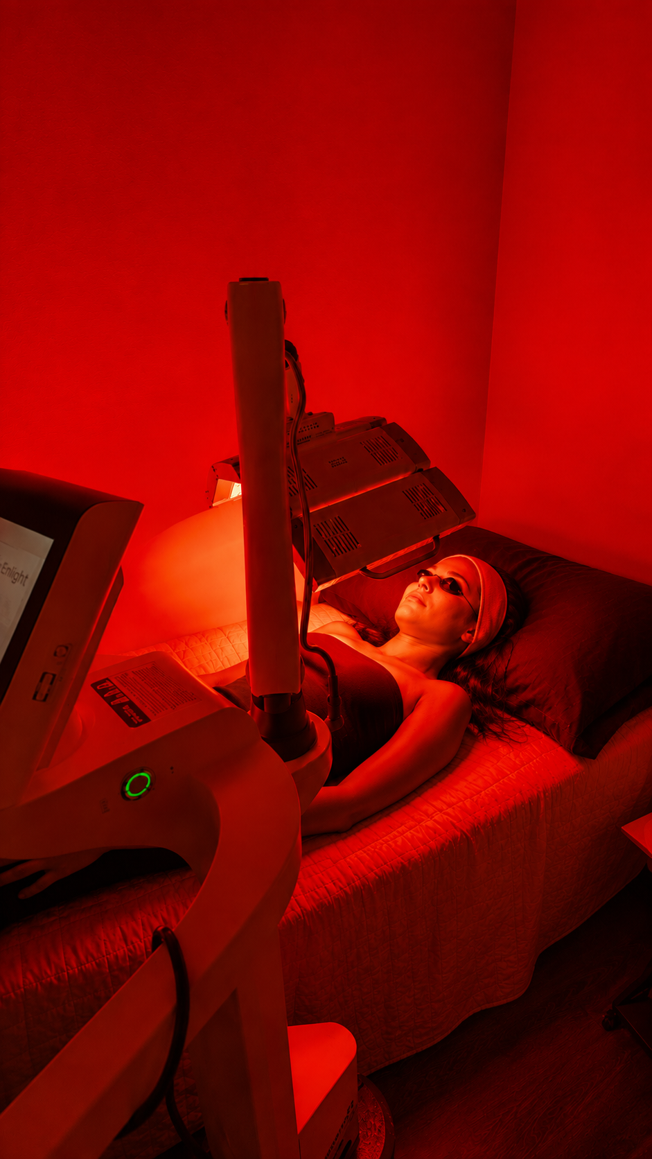 A woman lying on a bed undergoing laser skin treatment in a dark room with red lighting, wearing sunglasses and a black top, as a machine emits laser beams onto her face.