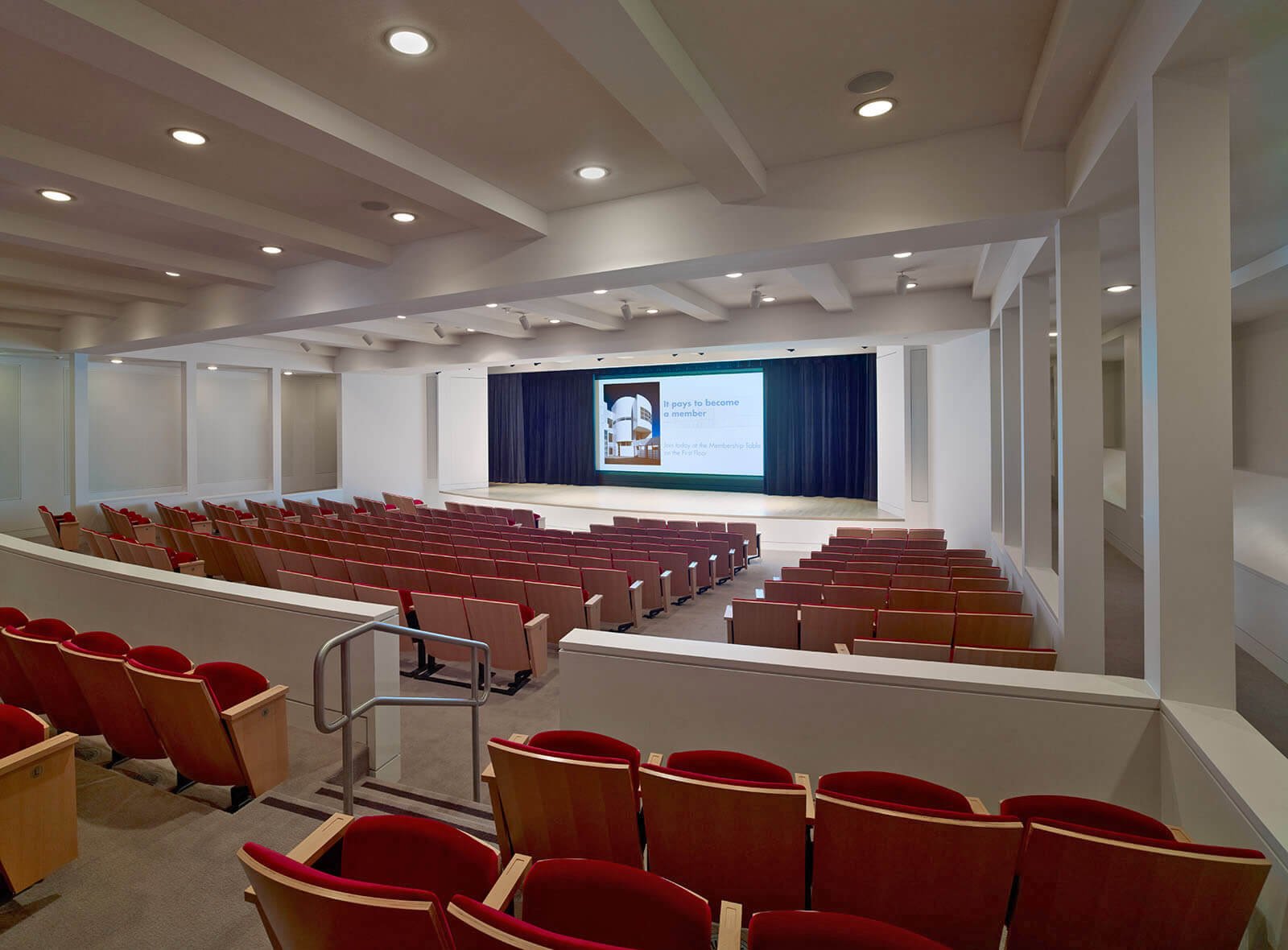 Empty auditorium with red chairs facing a stage with a large screen and blue curtains.