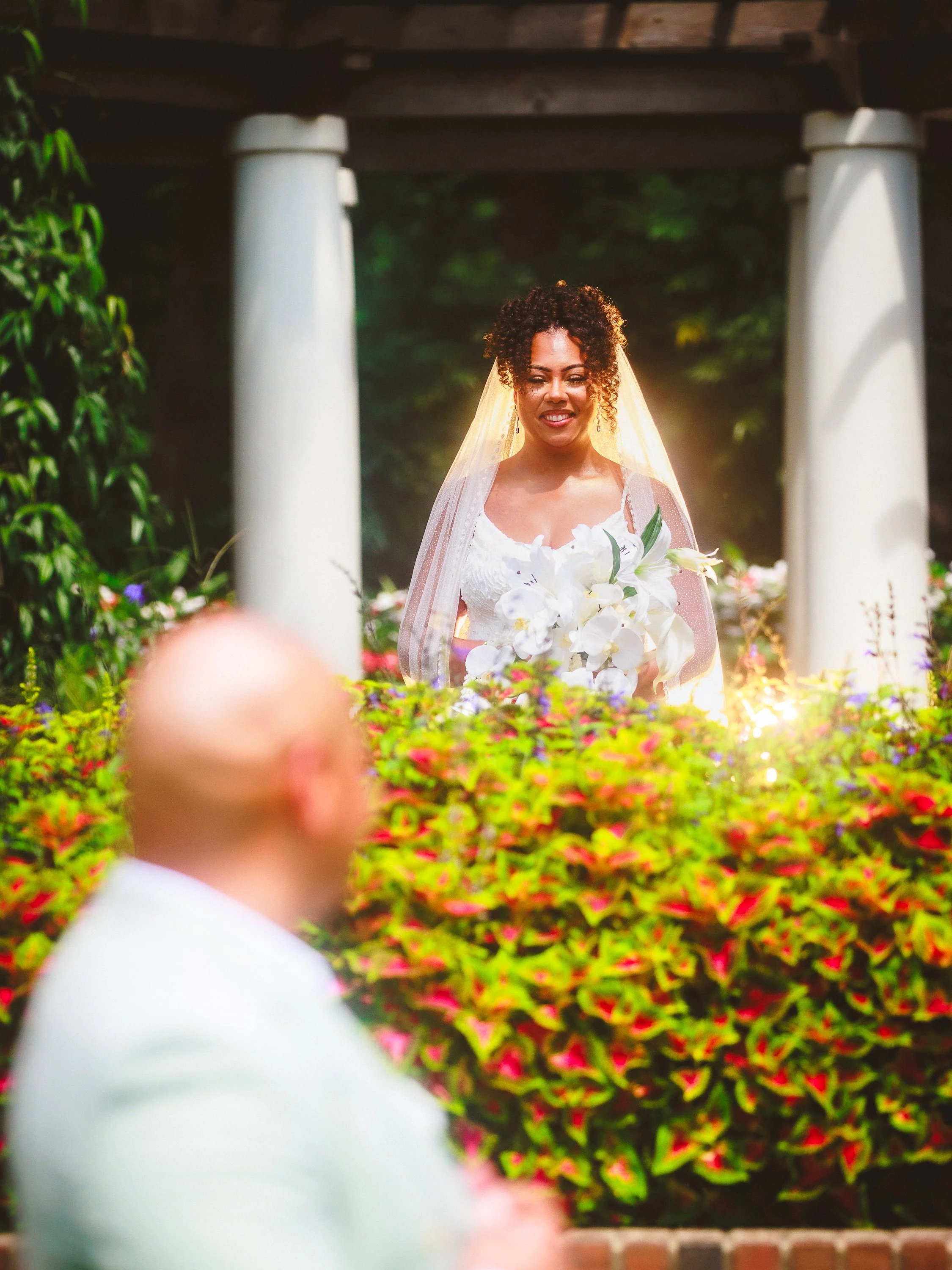 A bride holding a bouquet of white flowers enchanted by sunlight, standing amid colorful flowers near white columns, with a man in the foreground.