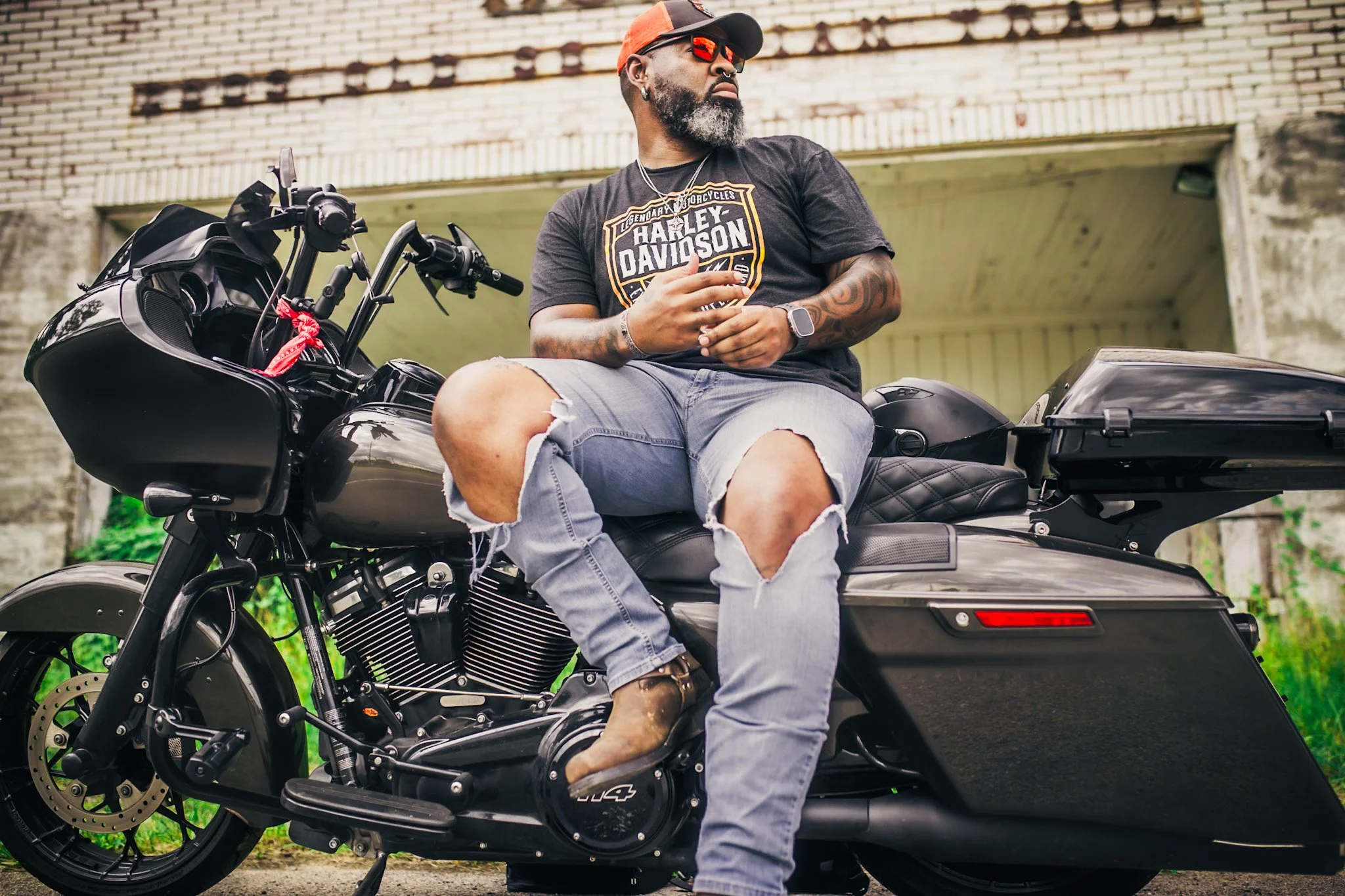 Man Pose Ideas for motorcycle . A bearded man wearing sunglasses, a Harley Davidson t-shirt, ripped jeans, and cowboy boots is sitting on a black motorcycle in front of an old brick building with a garage door.
