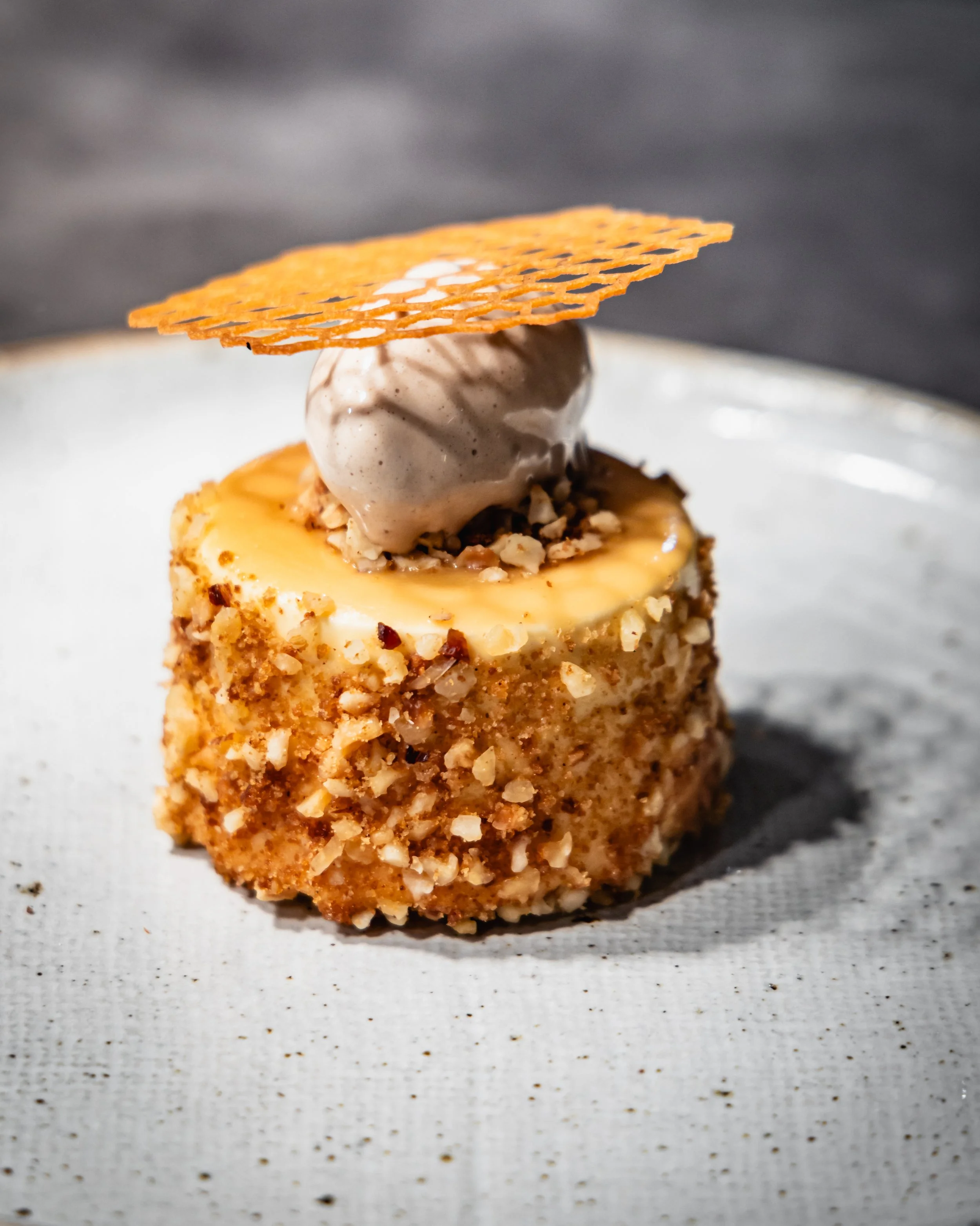 An elegant dessert featuring a round cake coated with chopped nuts, topped with a scoop of ice cream, and garnished with a thin, decorative caramel disk on top, served on a light-colored plate.