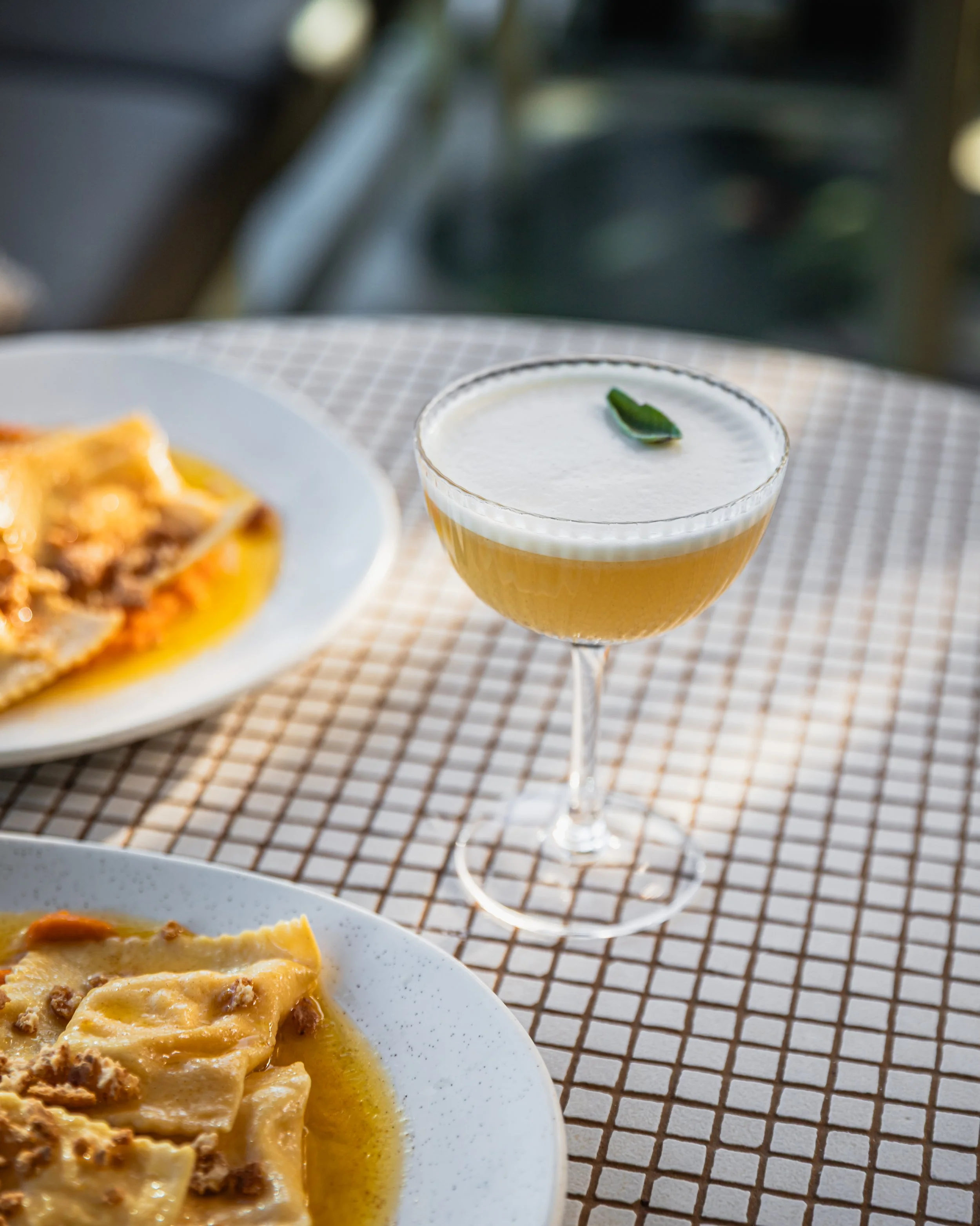 A cocktail glass with a lime wedge garnish on top, placed on a checkered tablecloth, beside plates of ravioli pasta with sauce.