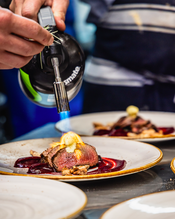 Chef torching a sliced cooked beef dish topped with a creamy sauce on a white plate with berry sauce underneath, in a professional kitchen or restaurant setting.