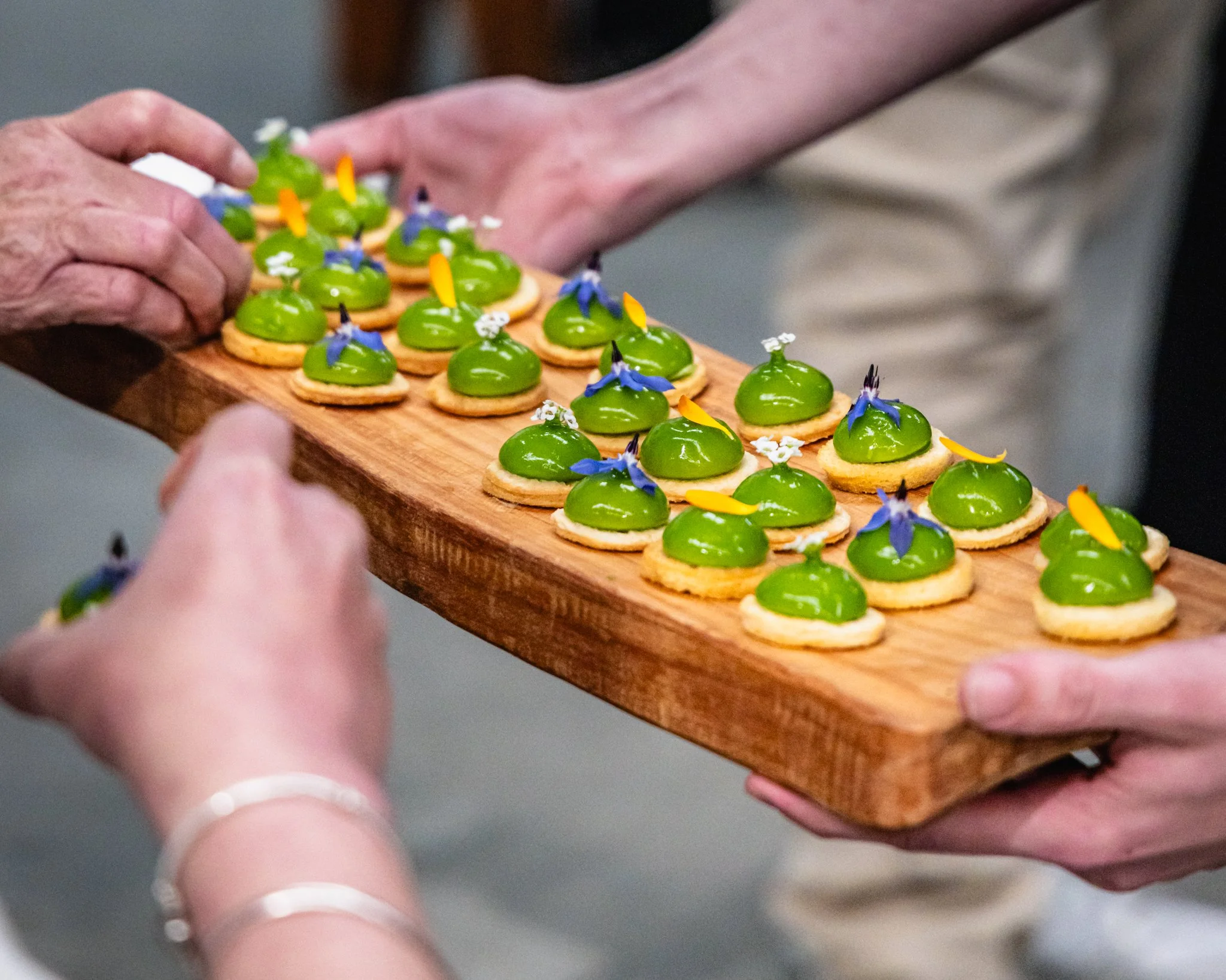 A person handing a wooden tray of small pastries topped with green glaze and edible flowers to another person.