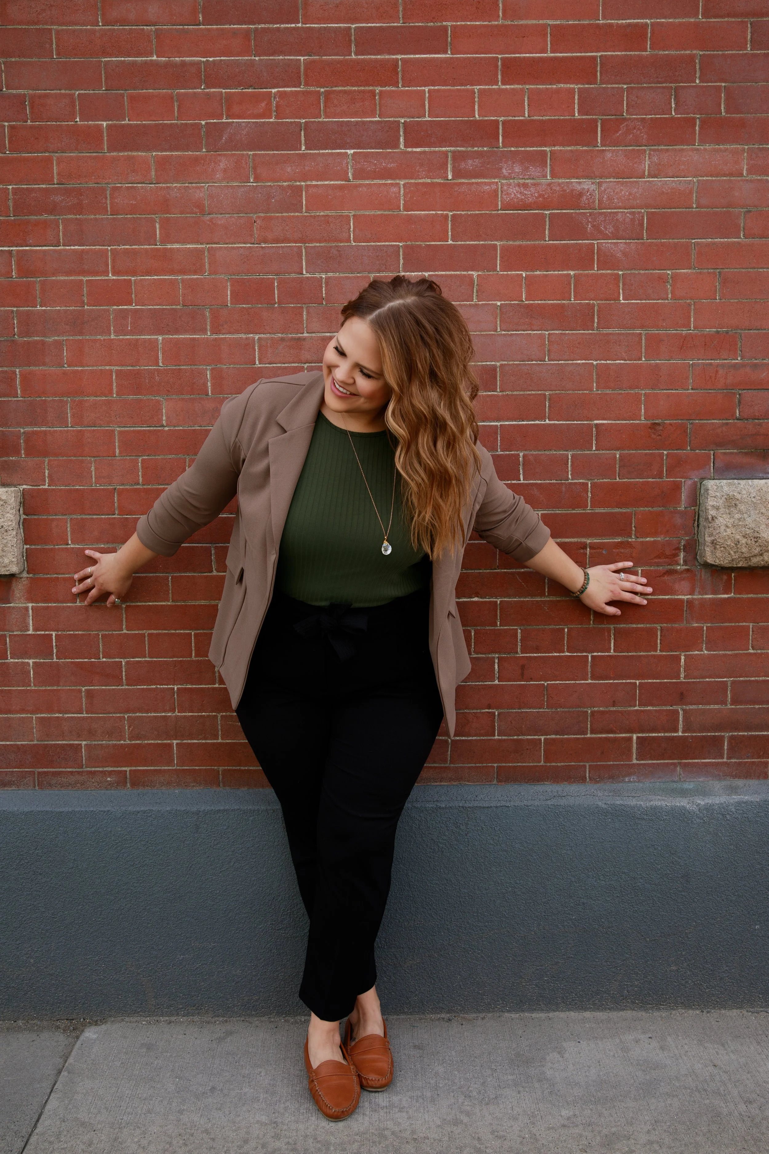 A woman with wavy, shoulder-length reddish-brown hair, wearing a green shirt, beige blazer, black pants, and brown loafers, standing against a red brick wall, smiling and looking down.
