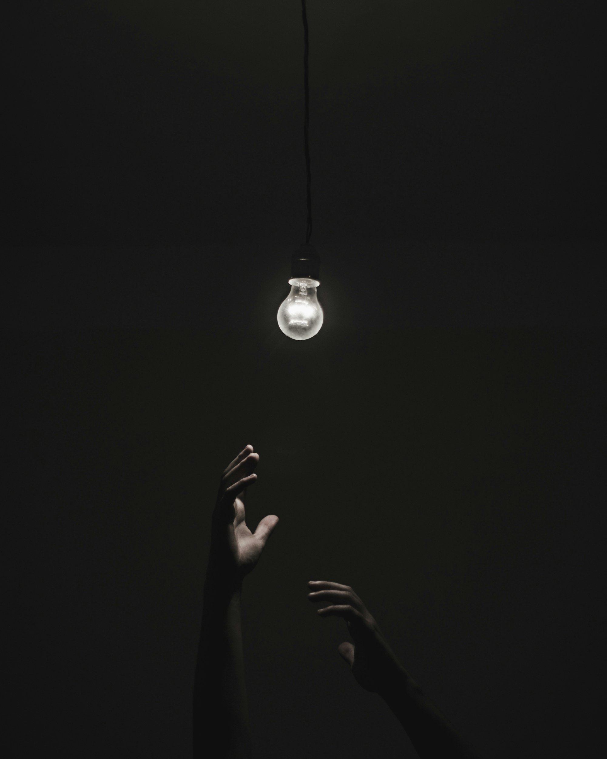 Dark room with a single hanging light bulb and two hands reaching upward beneath it.