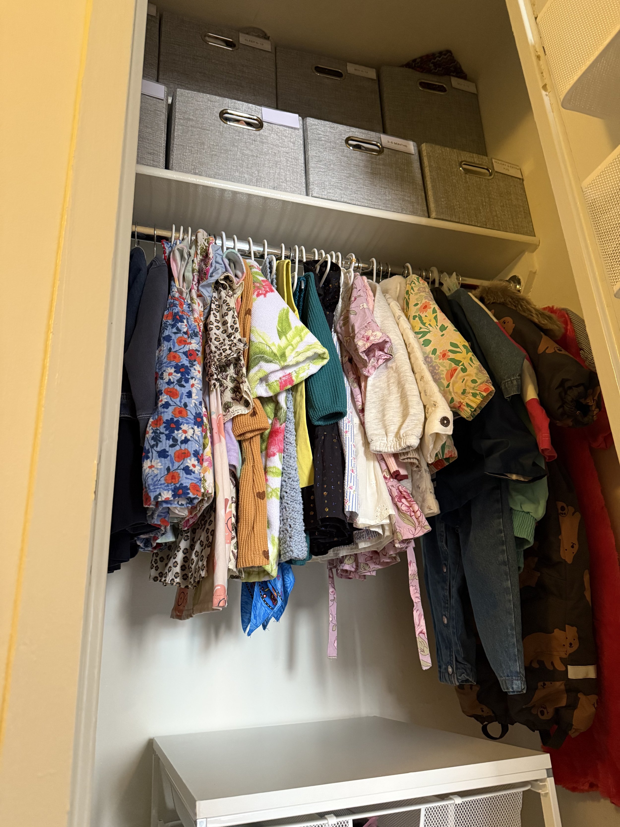 A closet with a row of colorful children's clothing hanging on a rod and storage boxes on a shelf above.