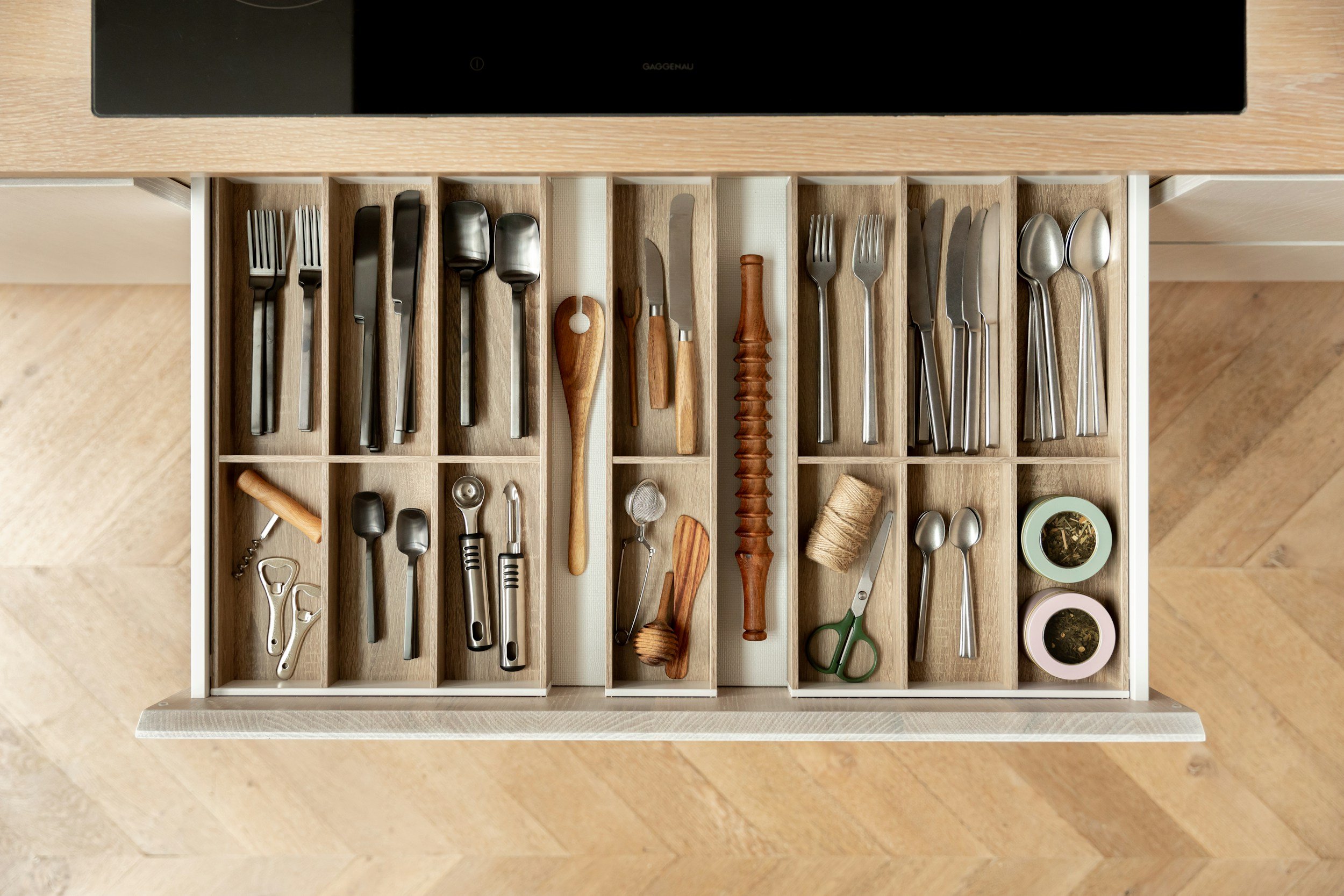 An open kitchen drawer filled with organized utensils and kitchen tools, including forks, knives, spoons, wooden spatulas, a corkscrew, scissors, tea strainers, and small containers of herbs.