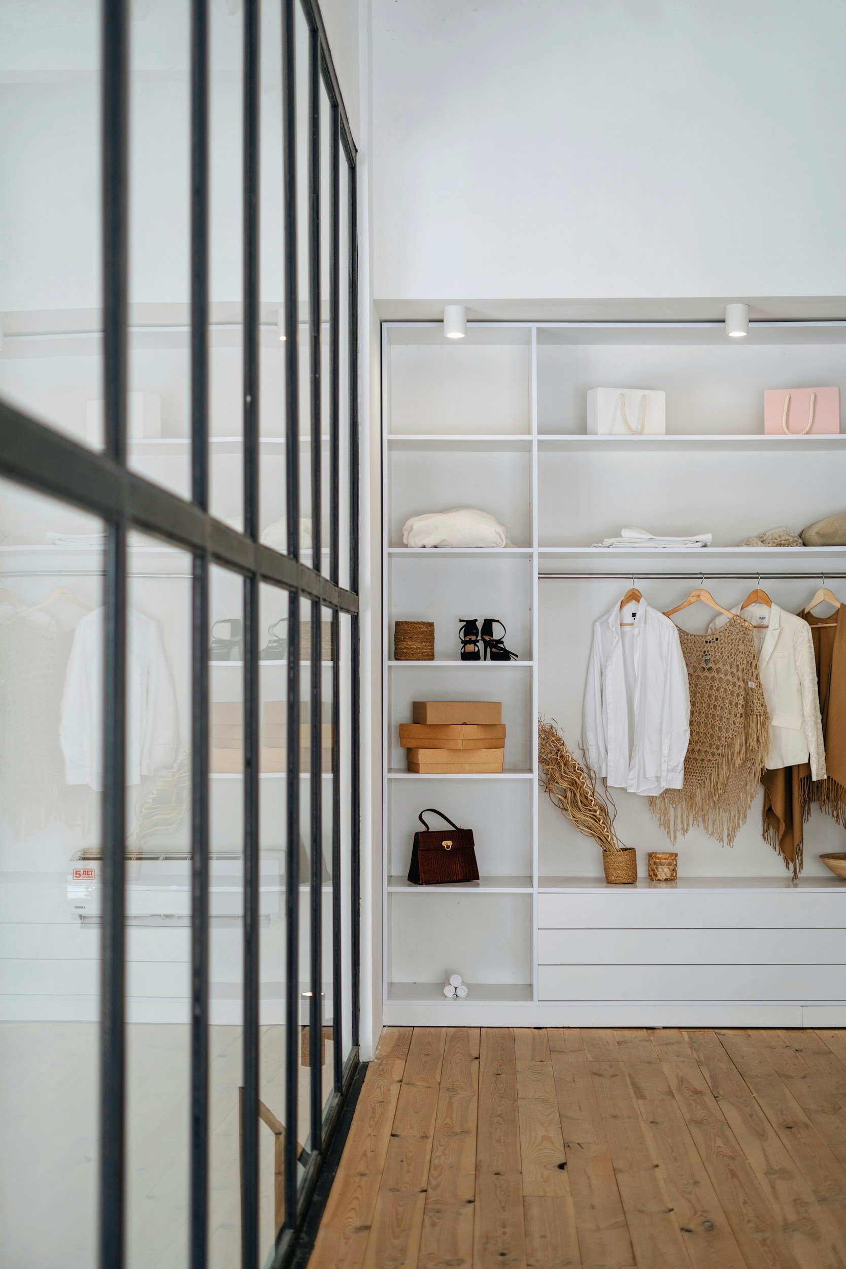 View of a white walk-in closet with shelves, hanging clothes, and decorative accessories, seen through a black metal-framed glass door.