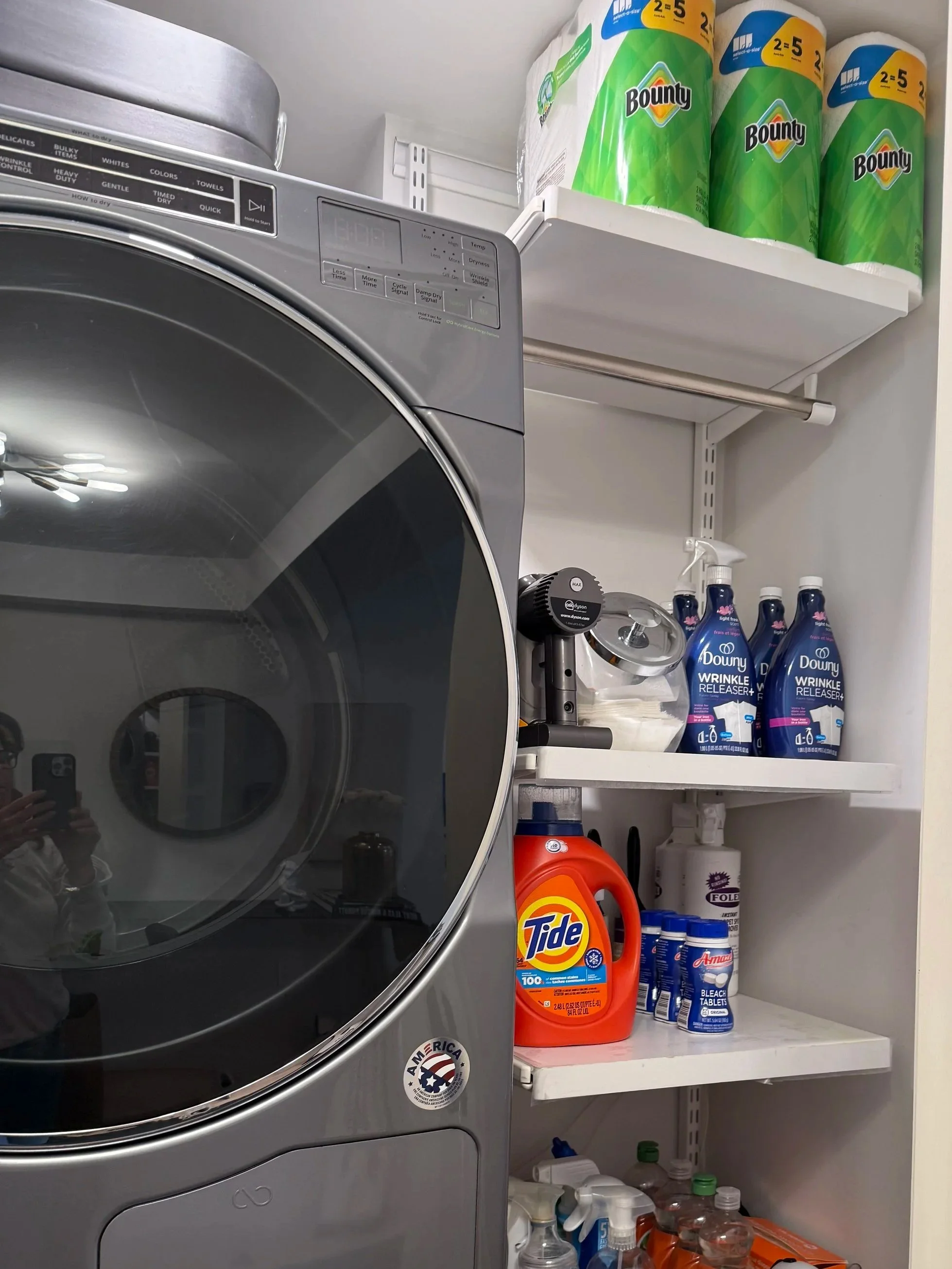 Laundry closet with a front-loading washing machine and shelves filled with cleaning supplies, paper towels, and laundry detergents.